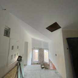 ZPRemBud Denys Shvets - Wnętrze w trakcie remontu: białe ściany, surowy beton na podłodze, widoczne okna i cegła. Na pierwszym planie lampa na statywie i płyta OSB.
