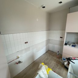 ZPRemBud Denys Shvets - Wnętrze kuchni w trakcie remontu: częściowo ułożone białe płytki na ścianie, widoczne instalacje, niezabudowane szafki i akcesoria remontowe na podłodze.