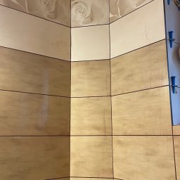 ZPRemBud Denys Shvets - Wykończona ściana w łazience w Piekarach Śląskich, z beżowymi płytkami z motywem kwiatowym. Widoczne fugi i fragment instalacji wodnej.