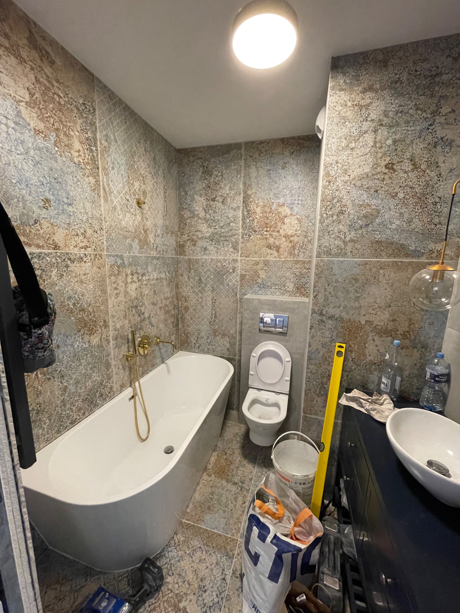 Ukończona łazienka z wanną, toaletą i umywalką. Na ścianach płytki imitujące wzór. Widoczne narzędzia i materiały budowlane sugerują prace wykończeniowe.