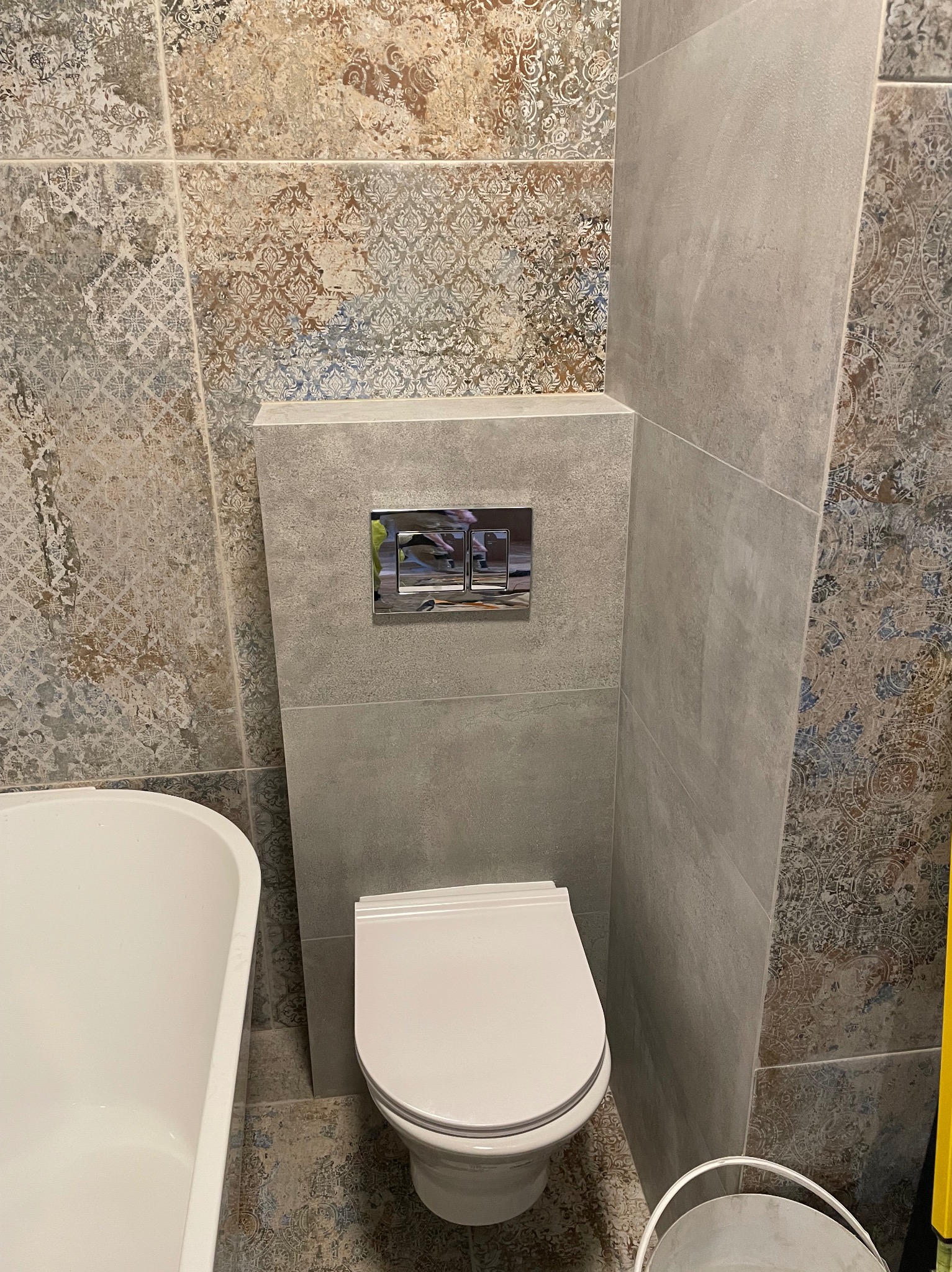 Wykończona łazienka: toaleta z chromowanym przyciskiem spłukiwania, ozdobne płytki w odcieniach beżu i szarości, fragment wanny. Nowoczesny design.