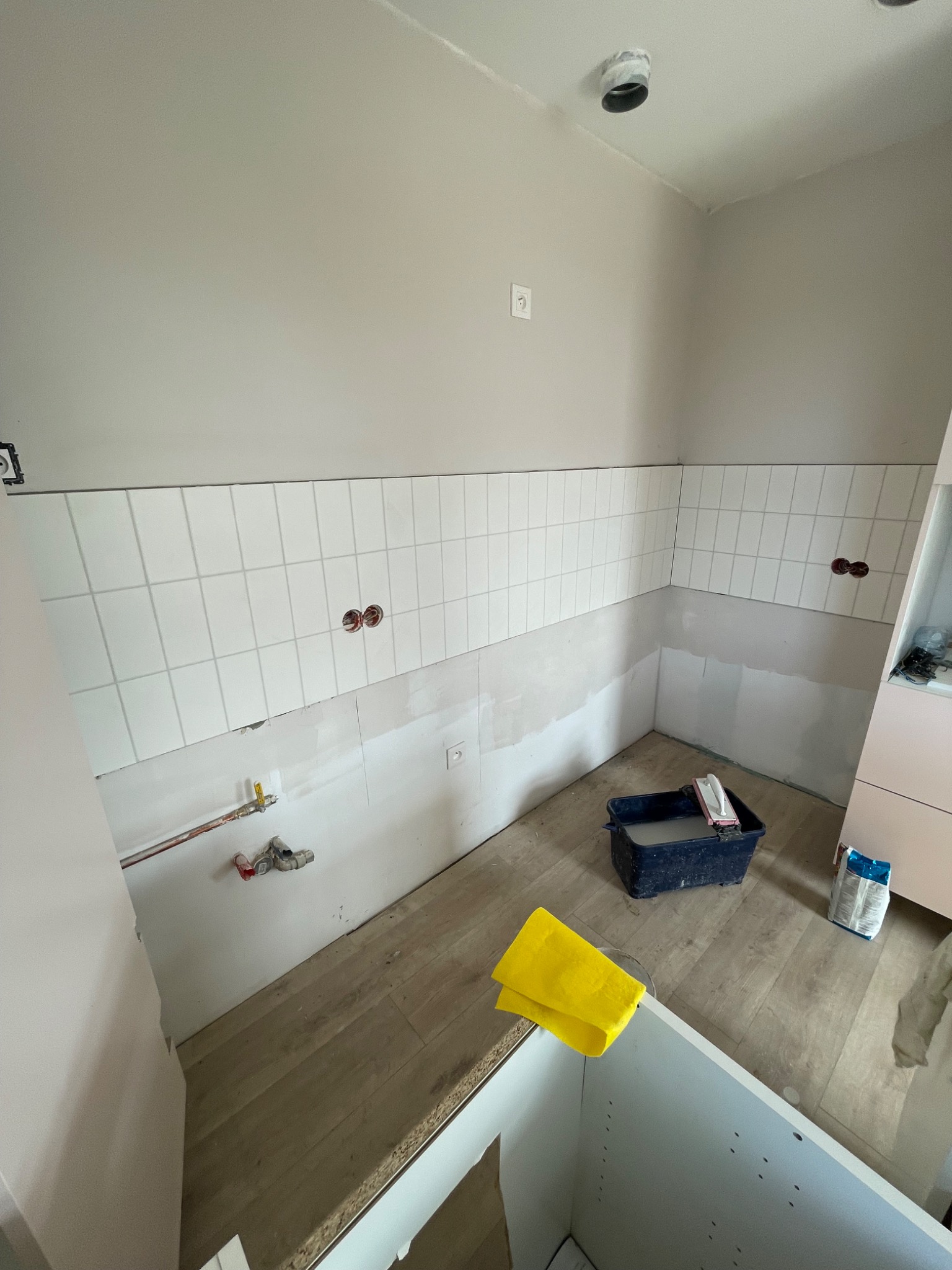 Remont kuchni w trakcie: białe płytki ułożone do połowy ściany, widoczne instalacje, wiadro z zaprawą i narzędzia. Podłoga wyłożona panelami imitującymi drewno.