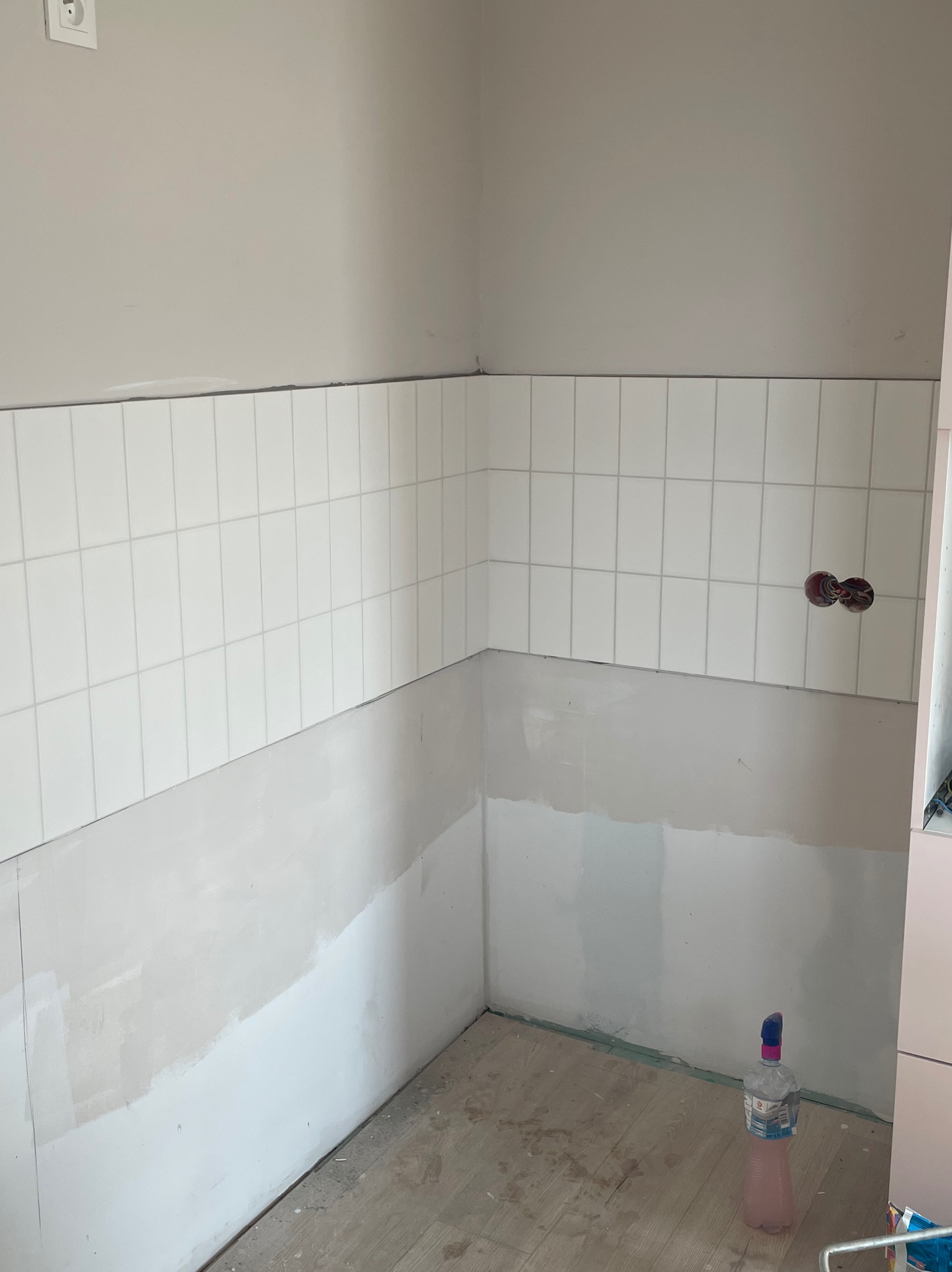 Białe, prostokątne płytki częściowo ułożone na ścianie w rogu pomieszczenia, dolna część ściany przygotowana do dalszych prac. Widoczne gniazdko elektryczne.