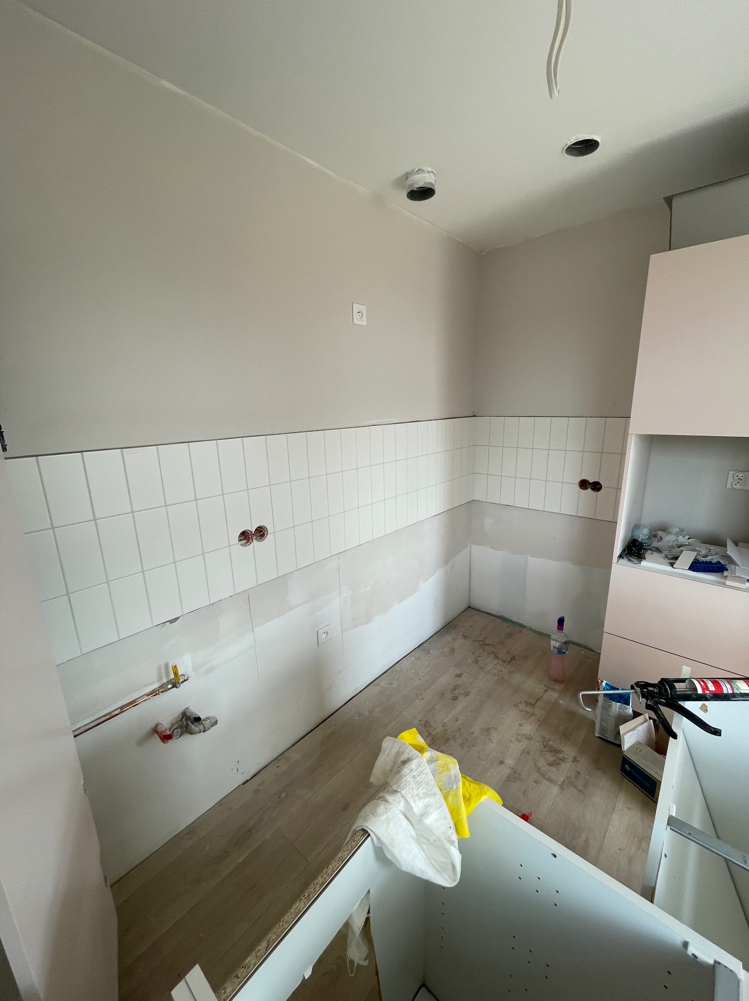 Białe płytki ułożone na ścianie w kuchni, widoczne instalacje i przygotowane miejsce pod zabudowę meblową. Częściowo wykończone wnętrze, stan surowy.