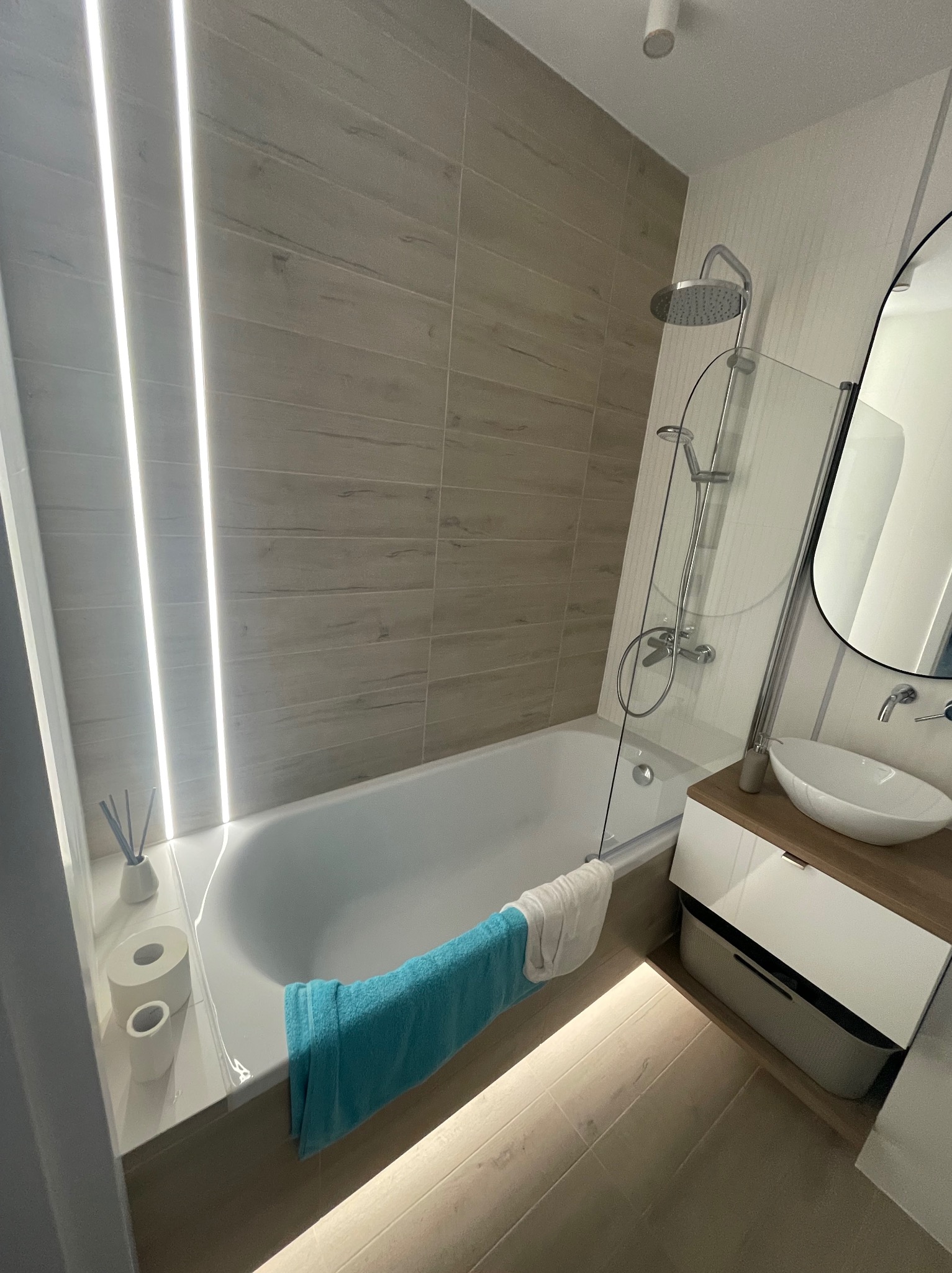 Nowoczesna łazienka z wanną, szklanym parawanem, umywalką na szafce i podświetleniem LED. Beżowe płytki imitujące drewno. Minimalistyczny design i jasne oświetlenie.