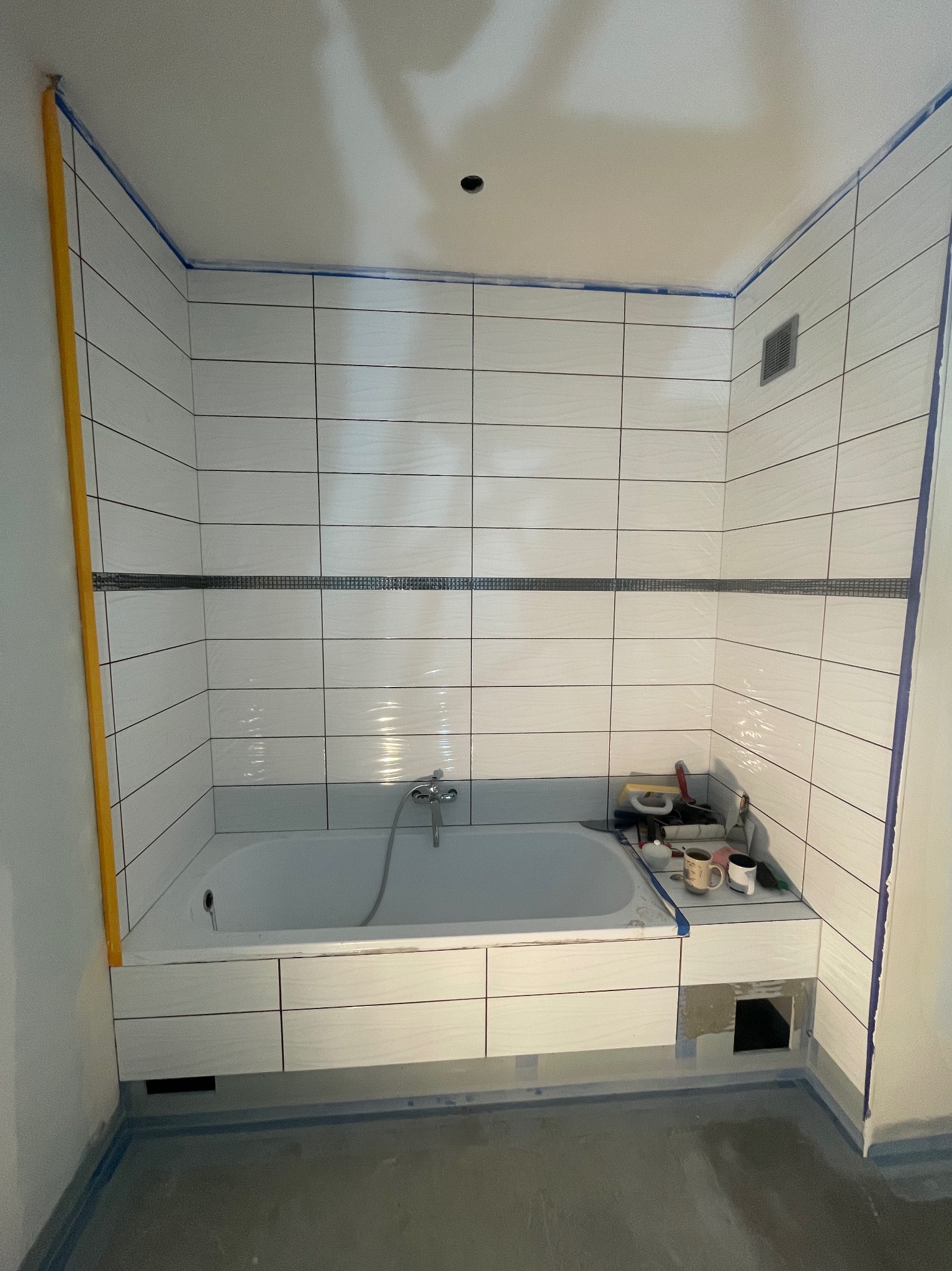 Biała wanna obudowana płytkami w łazience po częściowym remoncie. Widoczne narzędzia, poziomica, taśma malarska i fragment nieukończonej obudowy wanny.