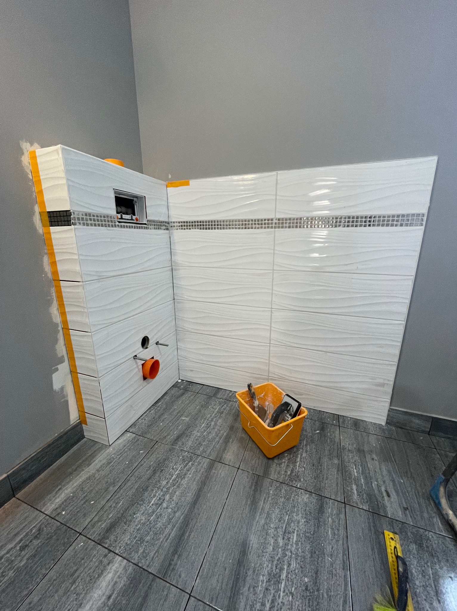 Białe, faliste płytki ścienne z dekoracyjnym pasem mozaiki, częściowo ułożone w rogu łazienki. Widoczne narzędzia glazurnicze w wiadrze. Szare płytki podłogowe.