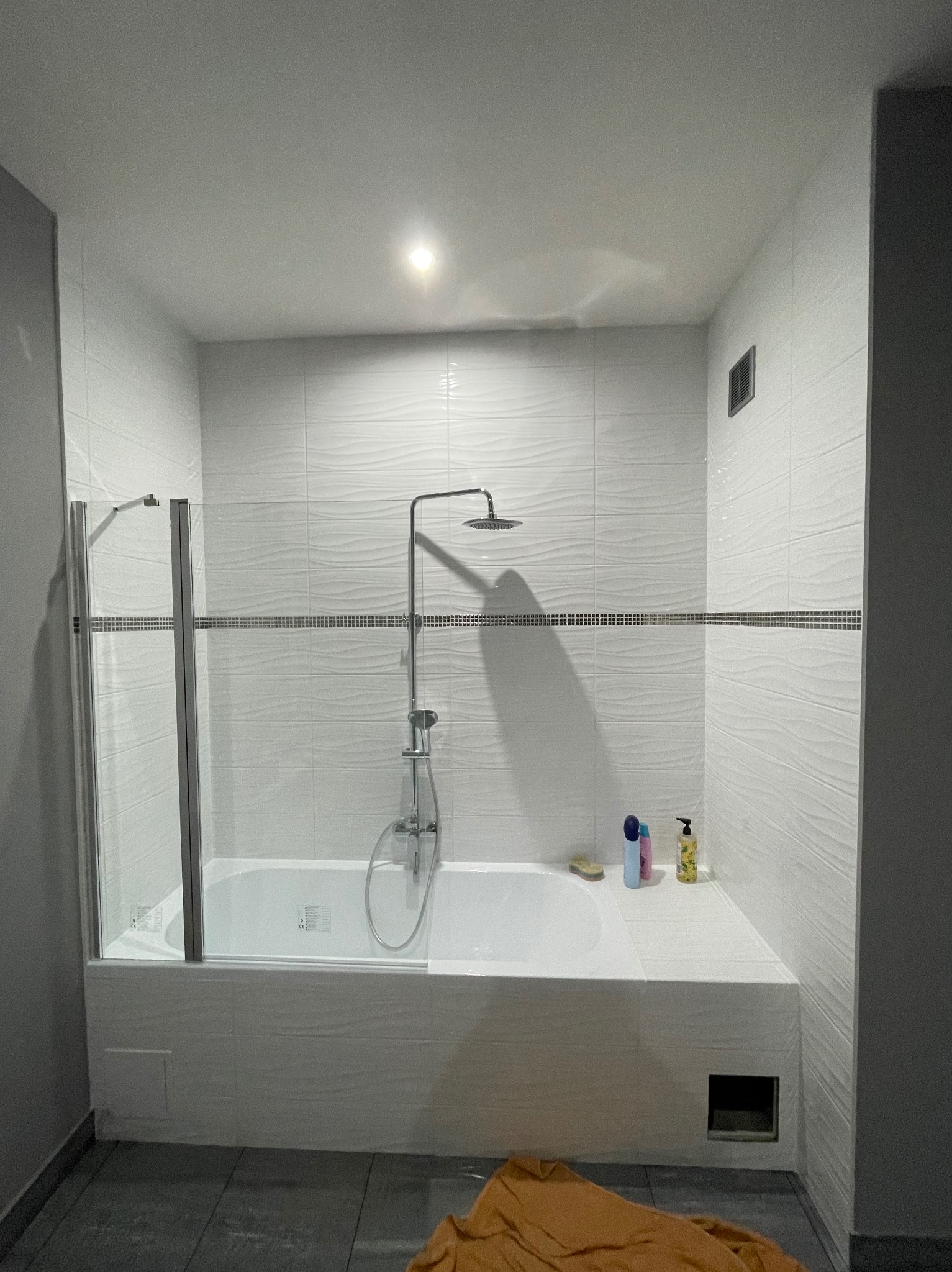 Nowoczesna łazienka z białą wanną obudowaną płytkami, szklanym parawanem i deszczownicą. Na wannie kosmetyki, na ścianach płytki z subtelną falistą strukturą.