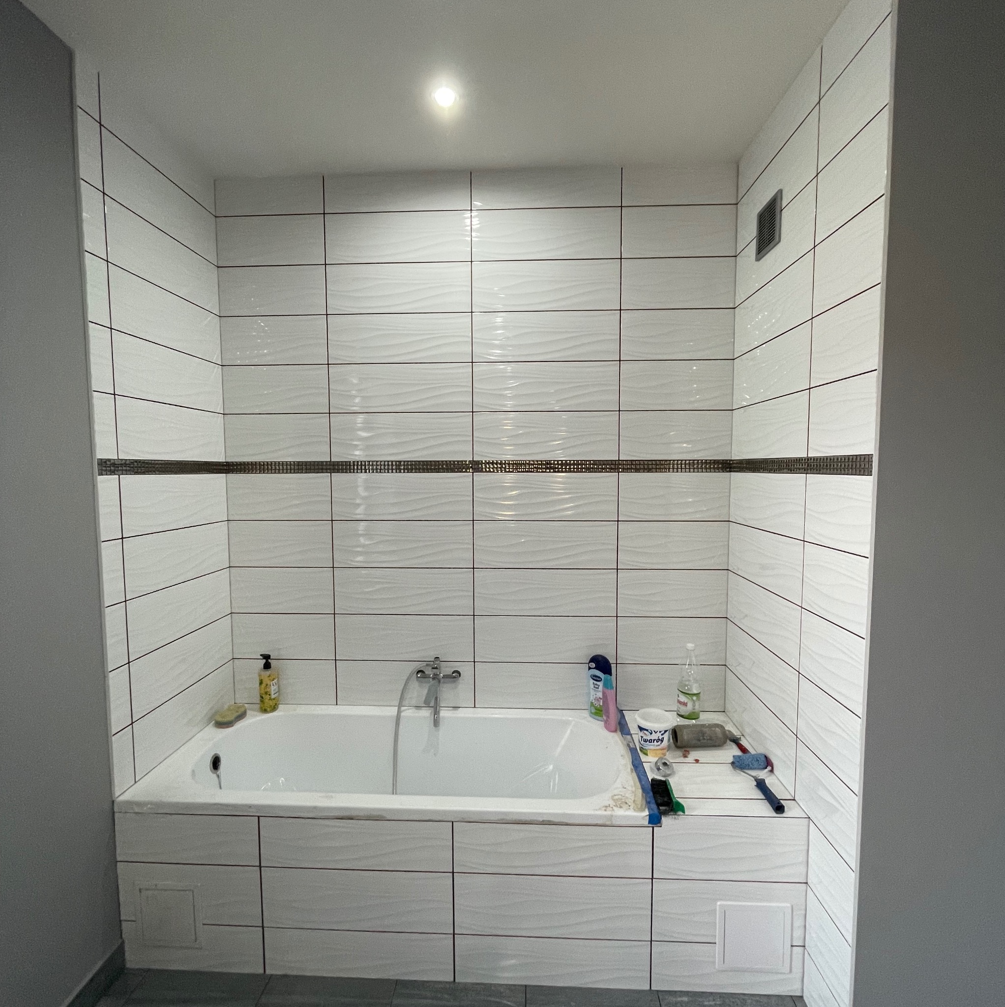 Wykończona łazienka z wanną obłożoną białymi, poziomymi płytkami z dekoracyjnym pasem. Widoczne akcesoria łazienkowe i narzędzia malarskie.