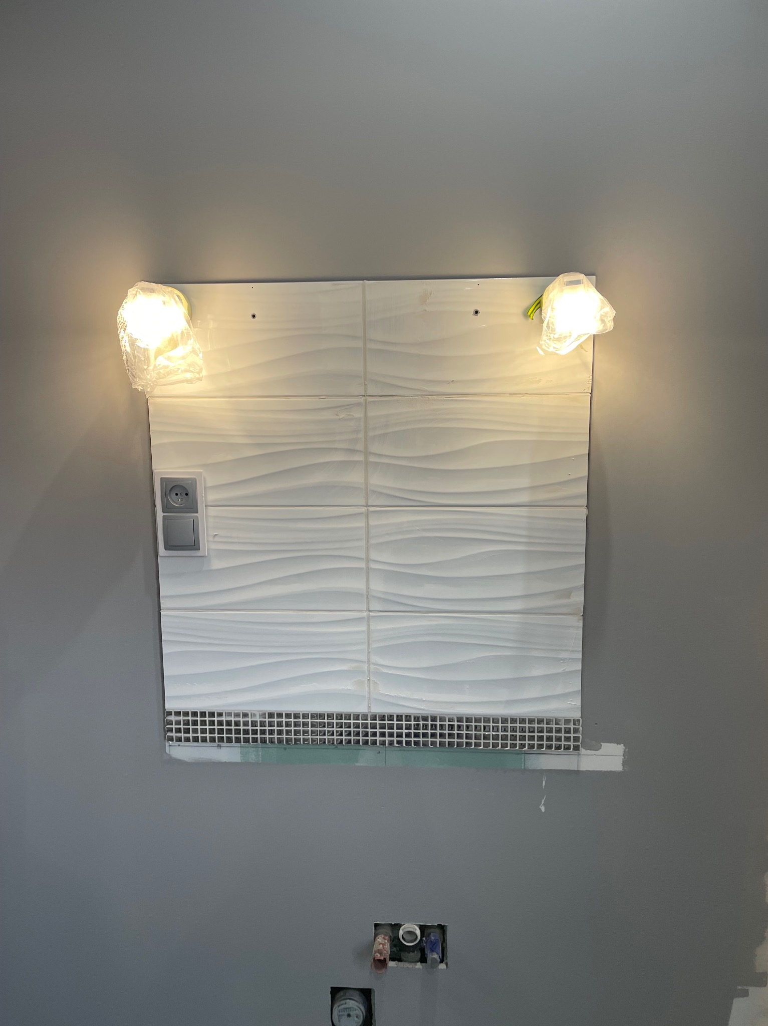 Ściana z falistych, białych płytek, z zamontowanym gniazdkiem i włącznikiem, podświetlona dwoma lampami w folii ochronnej, mozaika u dołu. Szare tło ściany.