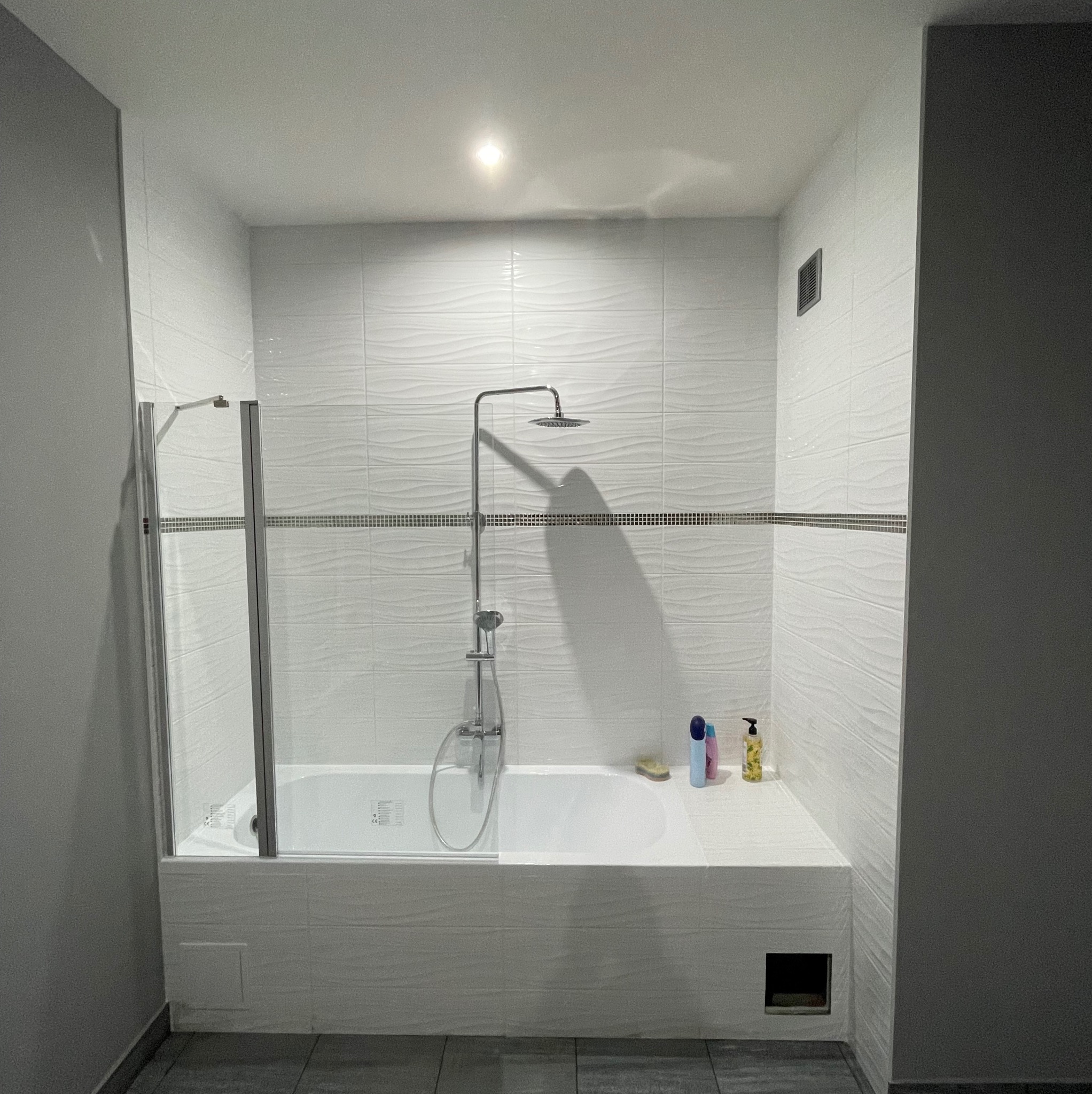 Biała łazienka z wanną i kabiną prysznicową, wykończona płytkami z subtelnym wzorem fali, akcentowana paskiem mozaiki. Widoczne kosmetyki i minimalistyczny design.