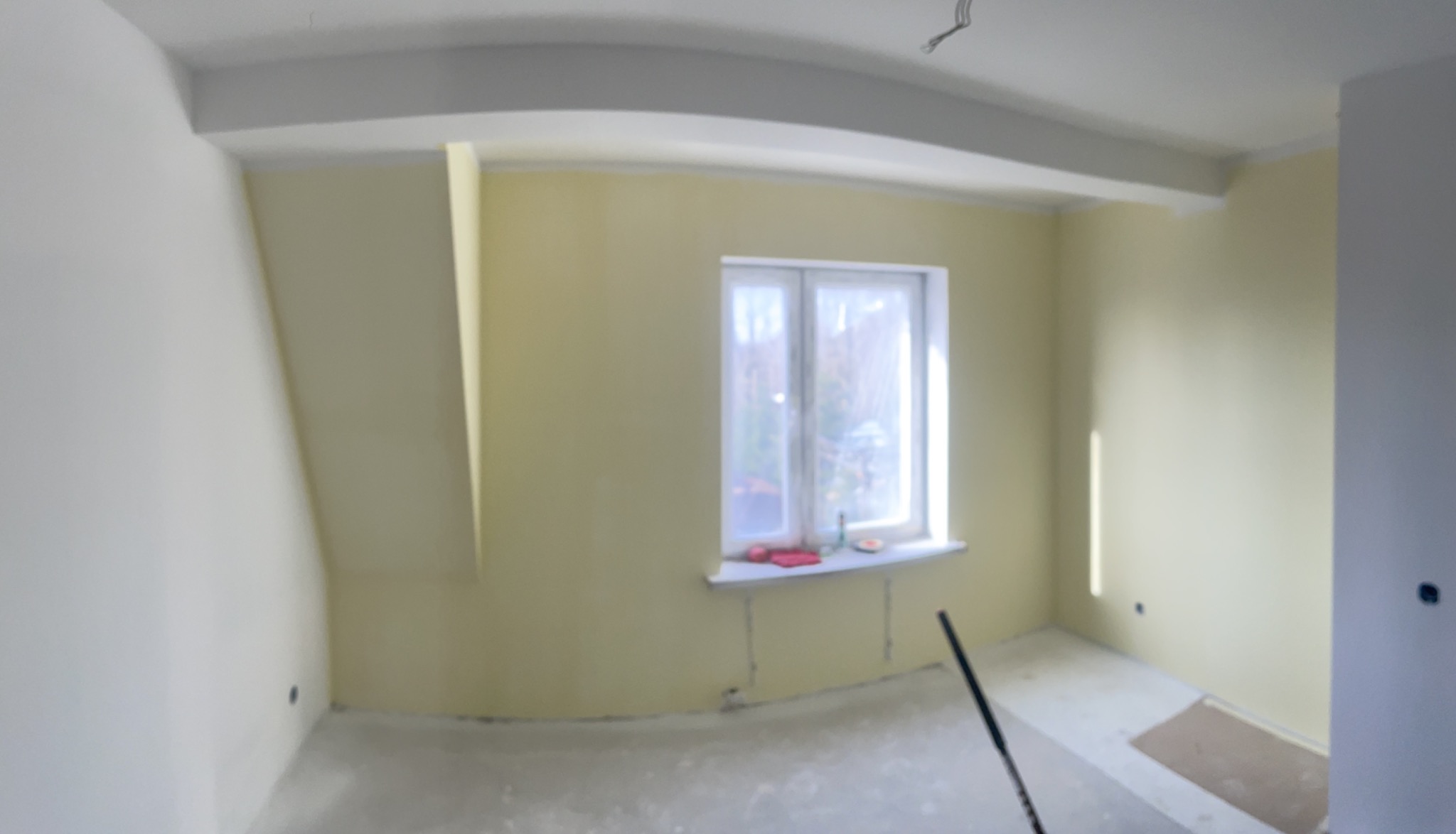 Pokój po malowaniu ścian na jasnożółty kolor, widoczne świeżo pomalowane ściany, okno z parapetem i fragment podłogi. W tle zarys drzew za oknem.
