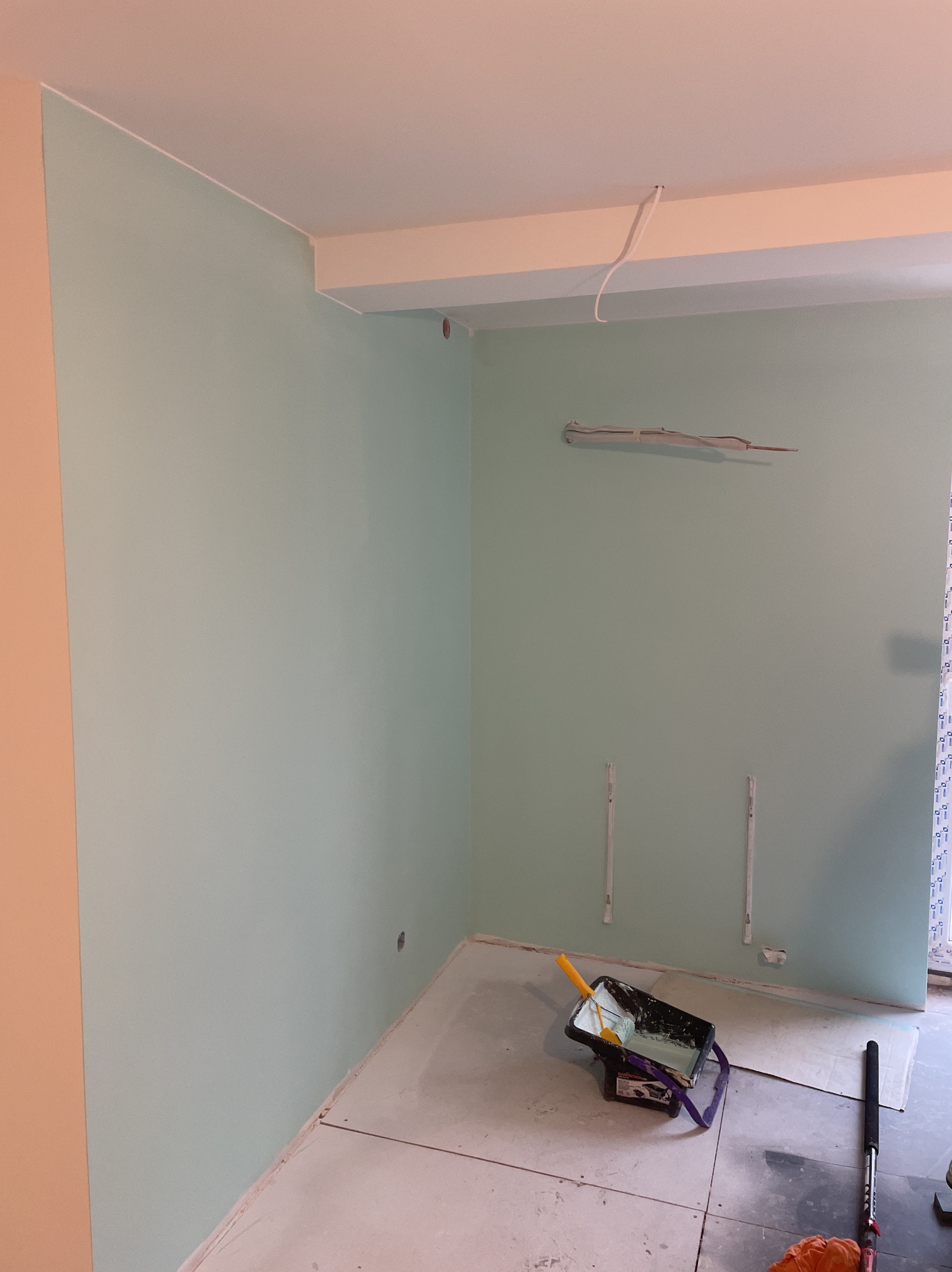 Świeżo pomalowane na jasnozielono ściany w rogu pokoju, widoczne narzędzia malarskie: kuweta z farbą i wałek. Podłoga przygotowana do dalszych prac wykończeniowych.