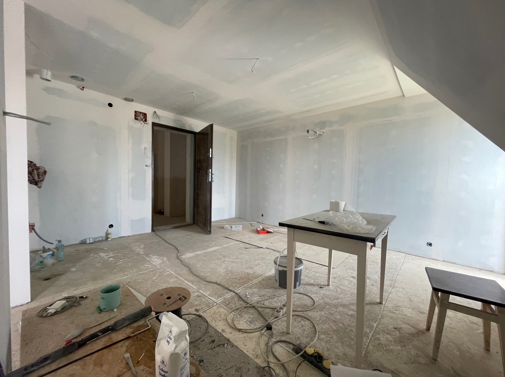 Pokój w trakcie remontu: ściany z płyt gipsowo-kartonowych, widoczne narzędzia i materiały, otwarte drzwi. Stolik z akcesoriami malarskimi w centrum kadru.