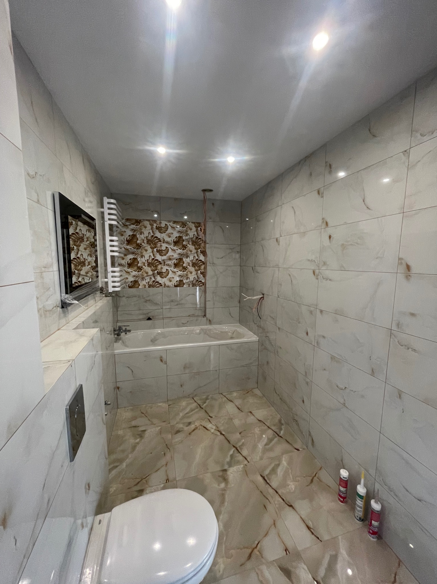 Jasna łazienka z marmurowymi płytkami, białą armaturą i grzejnikiem drabinkowym. Widoczne tuby z silikonem przy ścianie. Oświetlenie sufitowe punktowe.