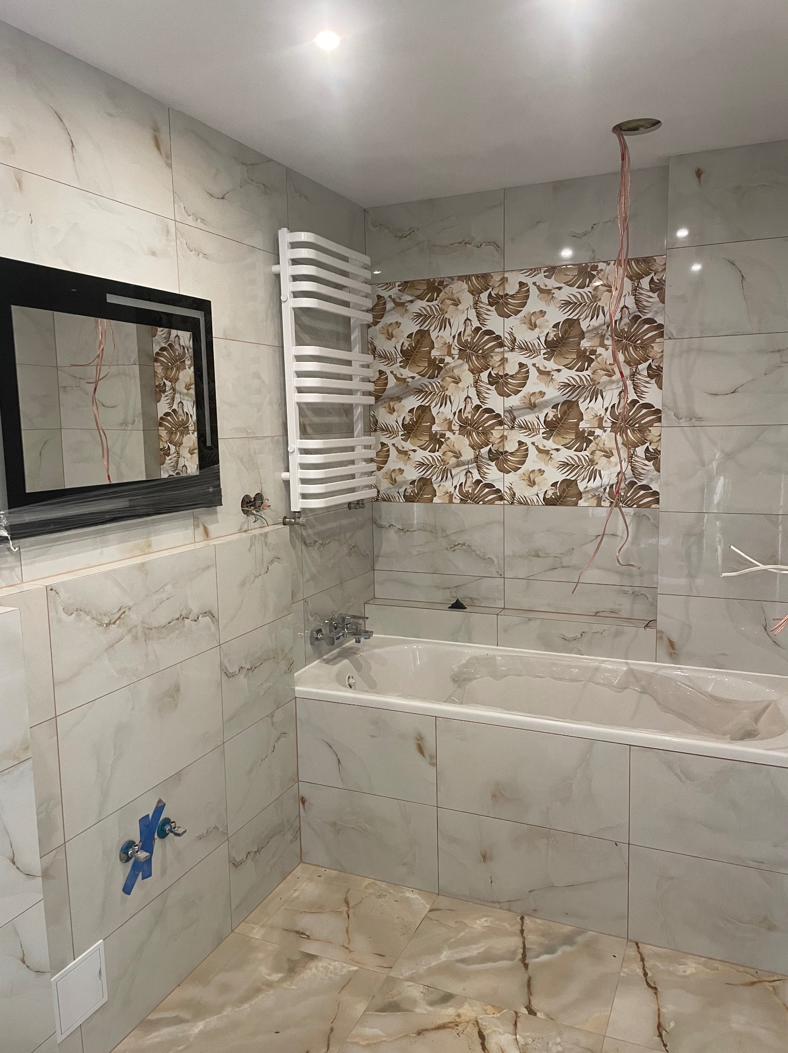 Wykończona łazienka w beżach: płytki imitujące marmur na ścianach i podłodze, wanna obudowana płytkami, grzejnik drabinkowy, lustro z czarną ramą, widoczne kable.