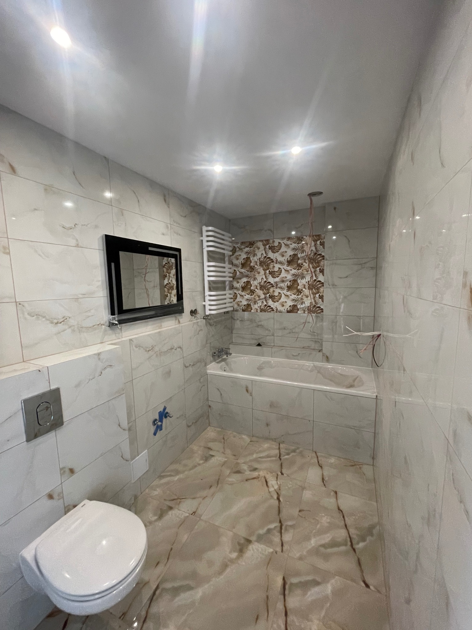 Wykończona łazienka z płytkami imitującymi marmur, wiszącą toaletą i czarnym lustrem. Fragmentalnie widoczne instalacje elektryczne.