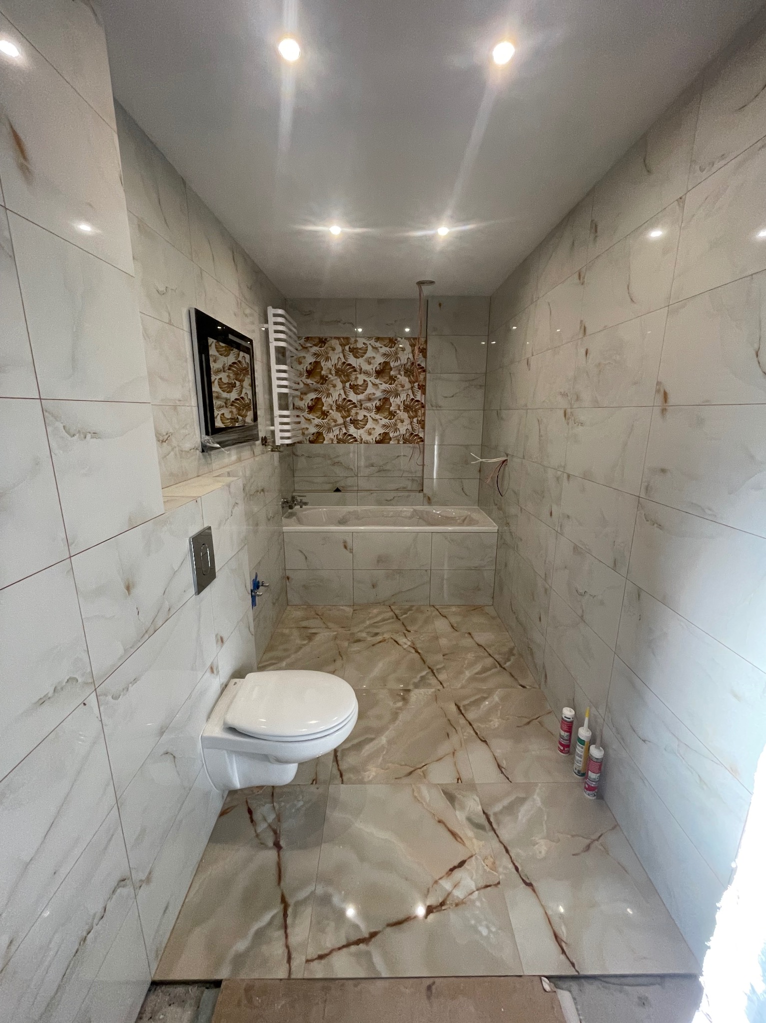 Nowoczesna łazienka po remoncie z białymi i brązowymi płytkami imitującymi marmur na ścianach i podłodze. Wisząca toaleta i grzejnik drabinkowy.