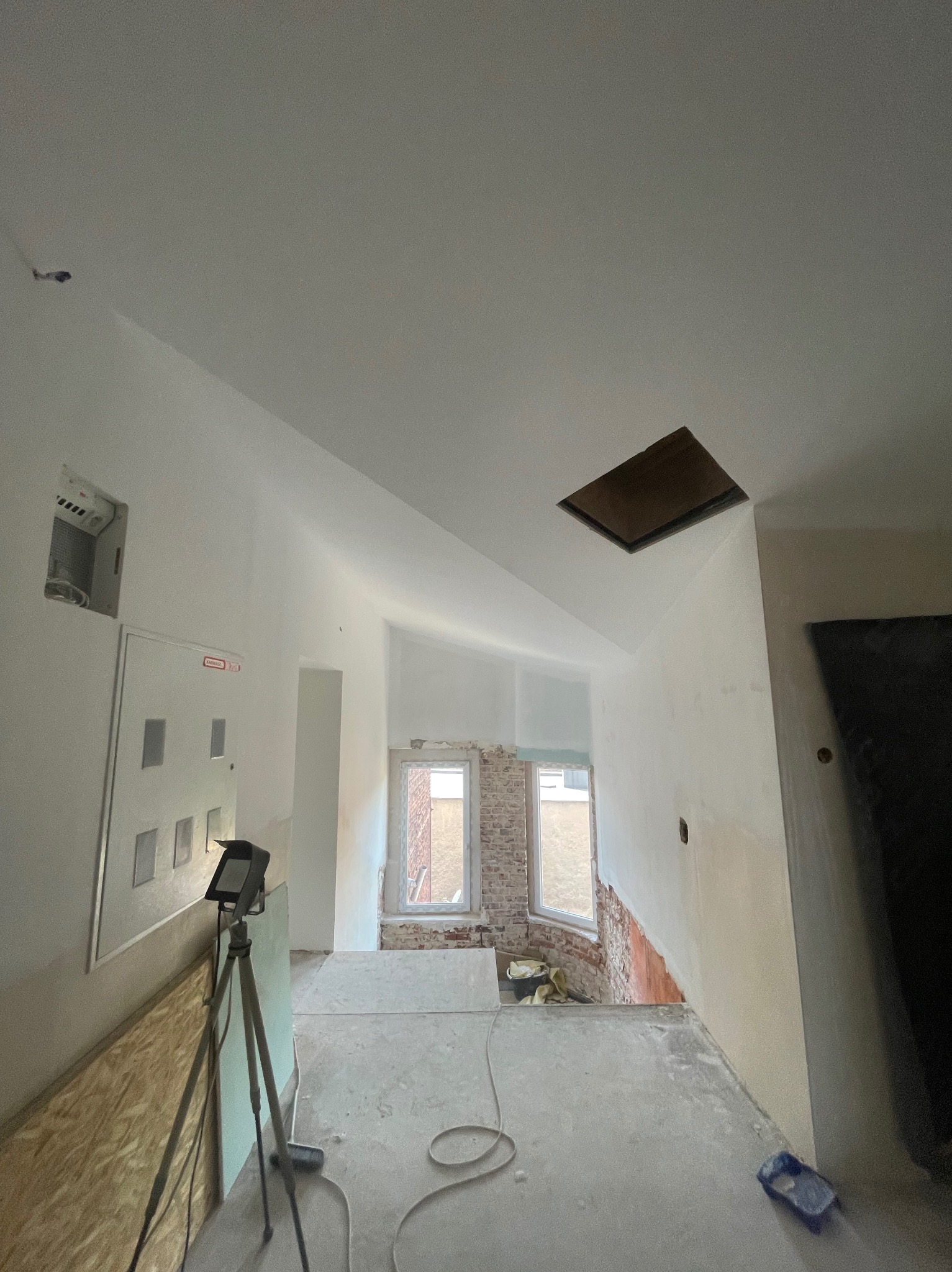 Wnętrze w trakcie remontu: białe ściany, surowy beton na podłodze, widoczne okna i cegła. Na pierwszym planie lampa na statywie i płyta OSB.