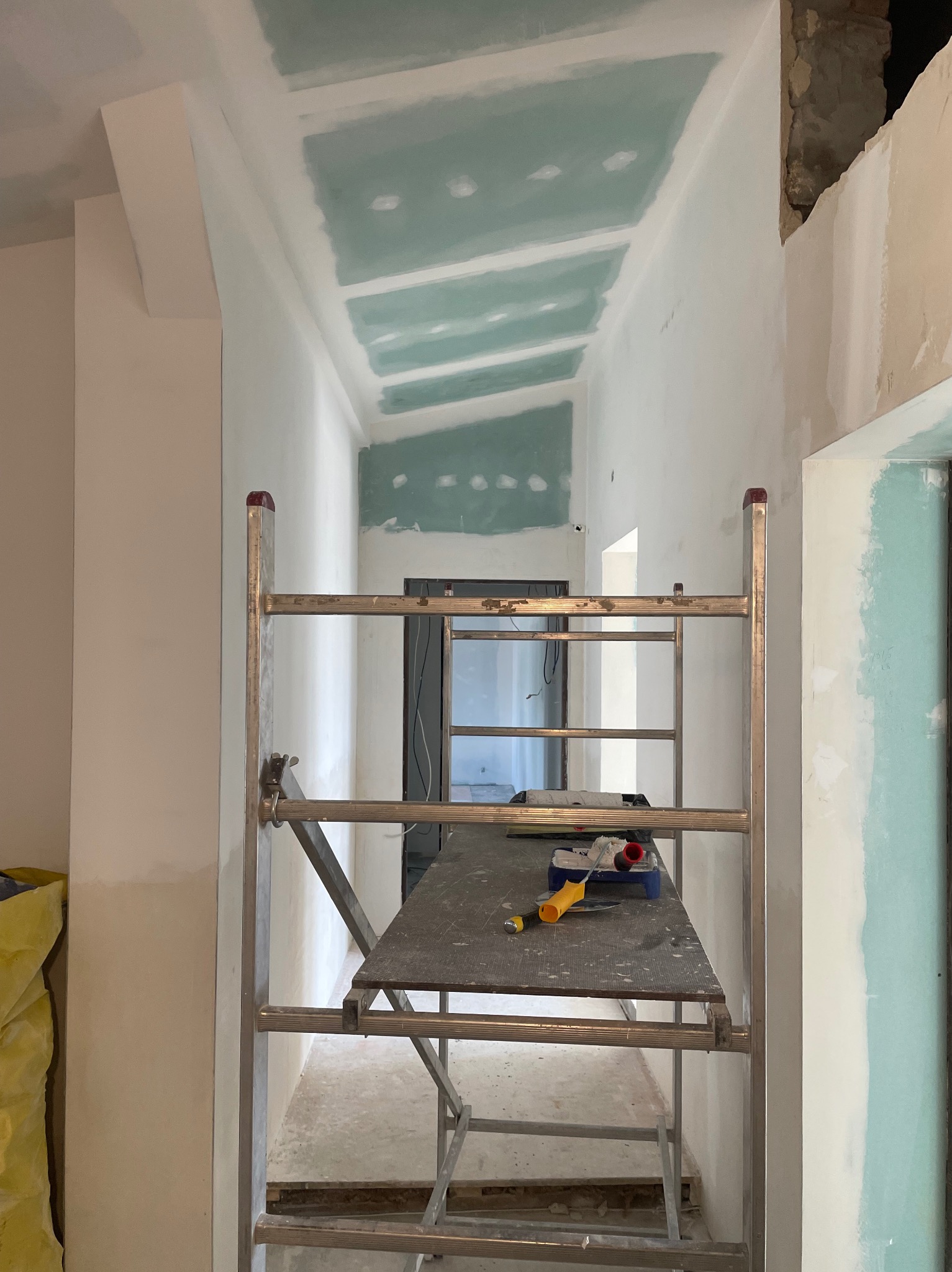 Wnętrze w trakcie prac: ściany i sufit pokryte częściowo zieloną warstwą, widoczne rusztowanie z narzędziami malarskimi. Fragment surowego muru.