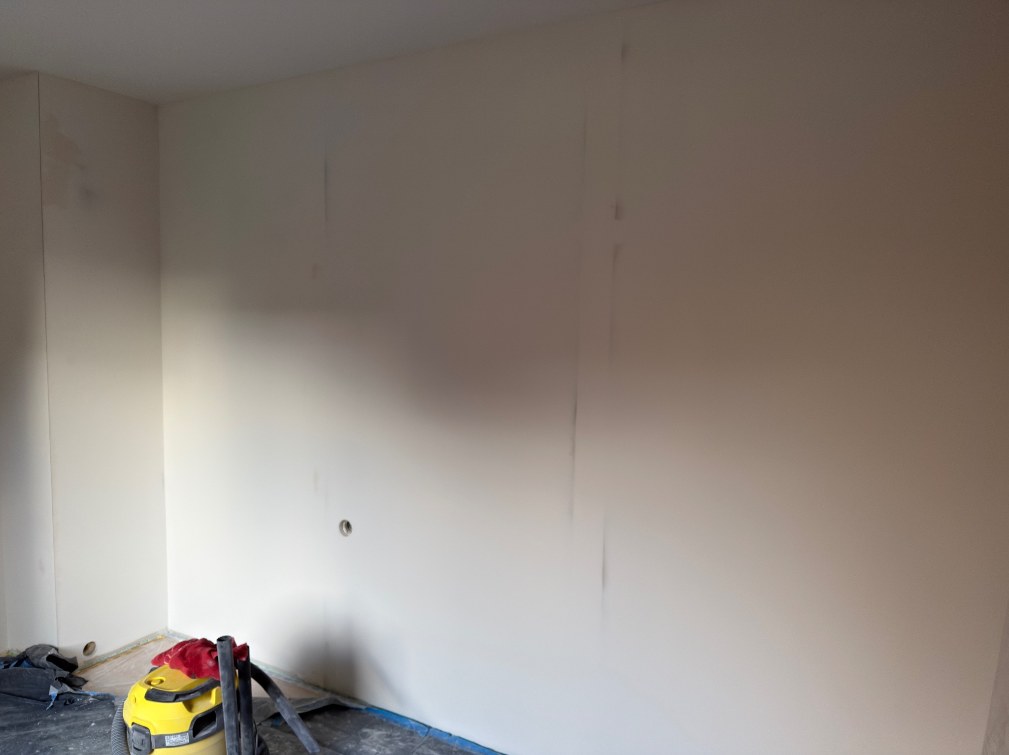 Wnętrze po szpachlowaniu ścian, widoczny żółty odkurzacz przemysłowy i zabezpieczona folią podłoga. Ściany przygotowane do dalszych prac wykończeniowych.