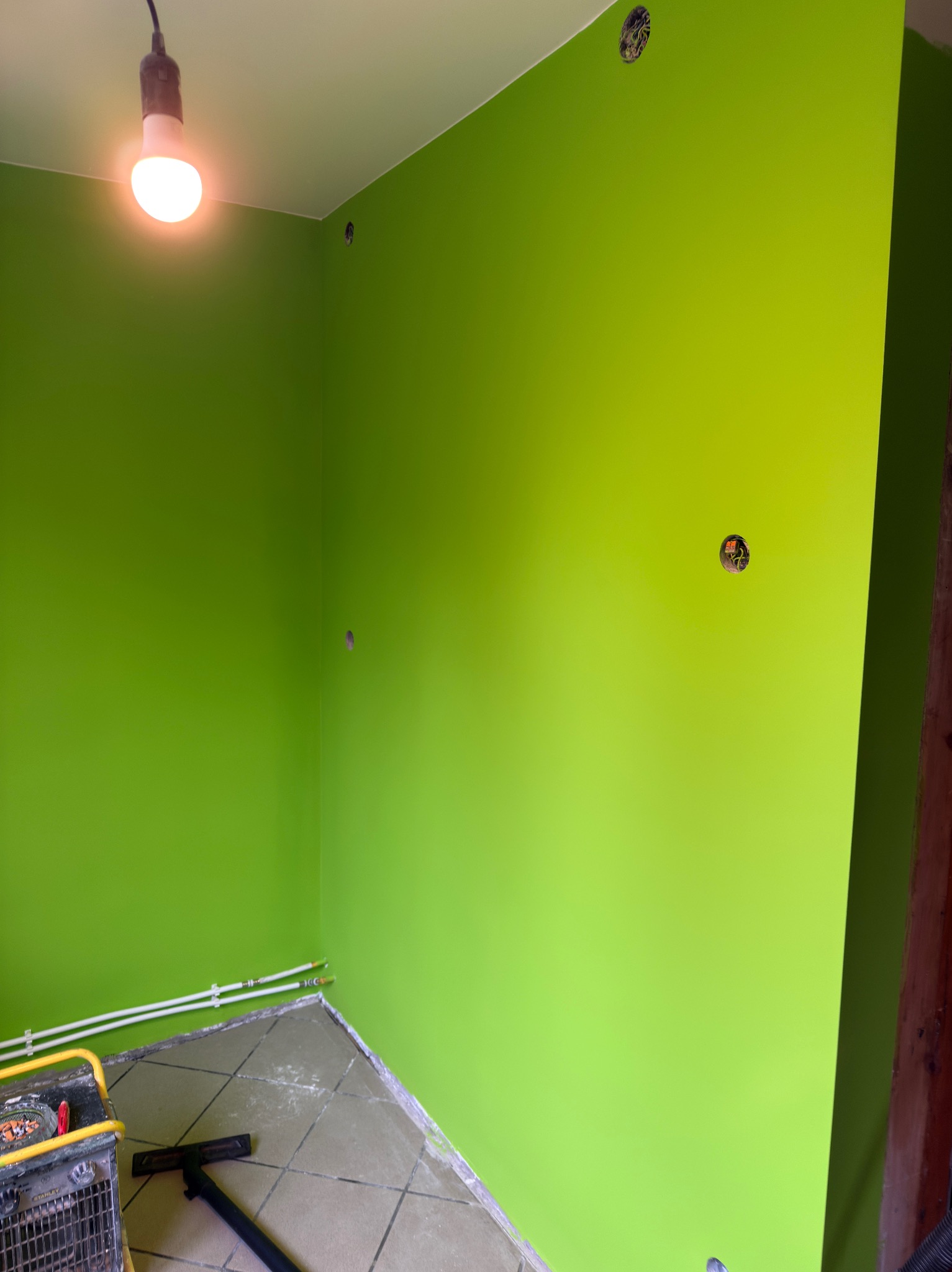 Świeżo pomalowane na jaskrawy zielony kolor ściany w rogu pokoju z widocznymi gniazdkami elektrycznymi i fragmentem podłogi z terakoty. Widoczna lampa i narzędzia.