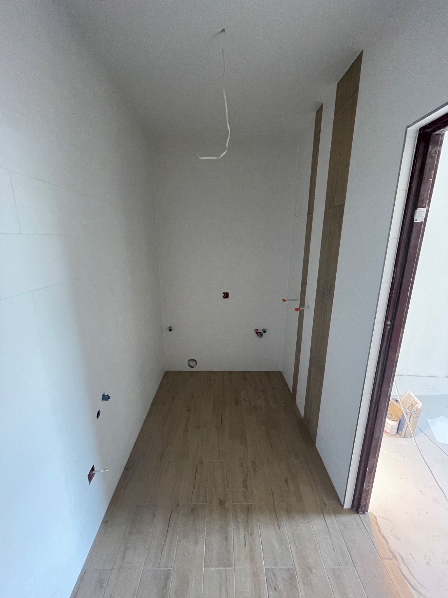 Wnętrze łazienki w trakcie wykańczania: ułożona podłoga z płytek imitujących drewno, częściowo wykończone ściany z widocznymi instalacjami i fragmentem drzwi.