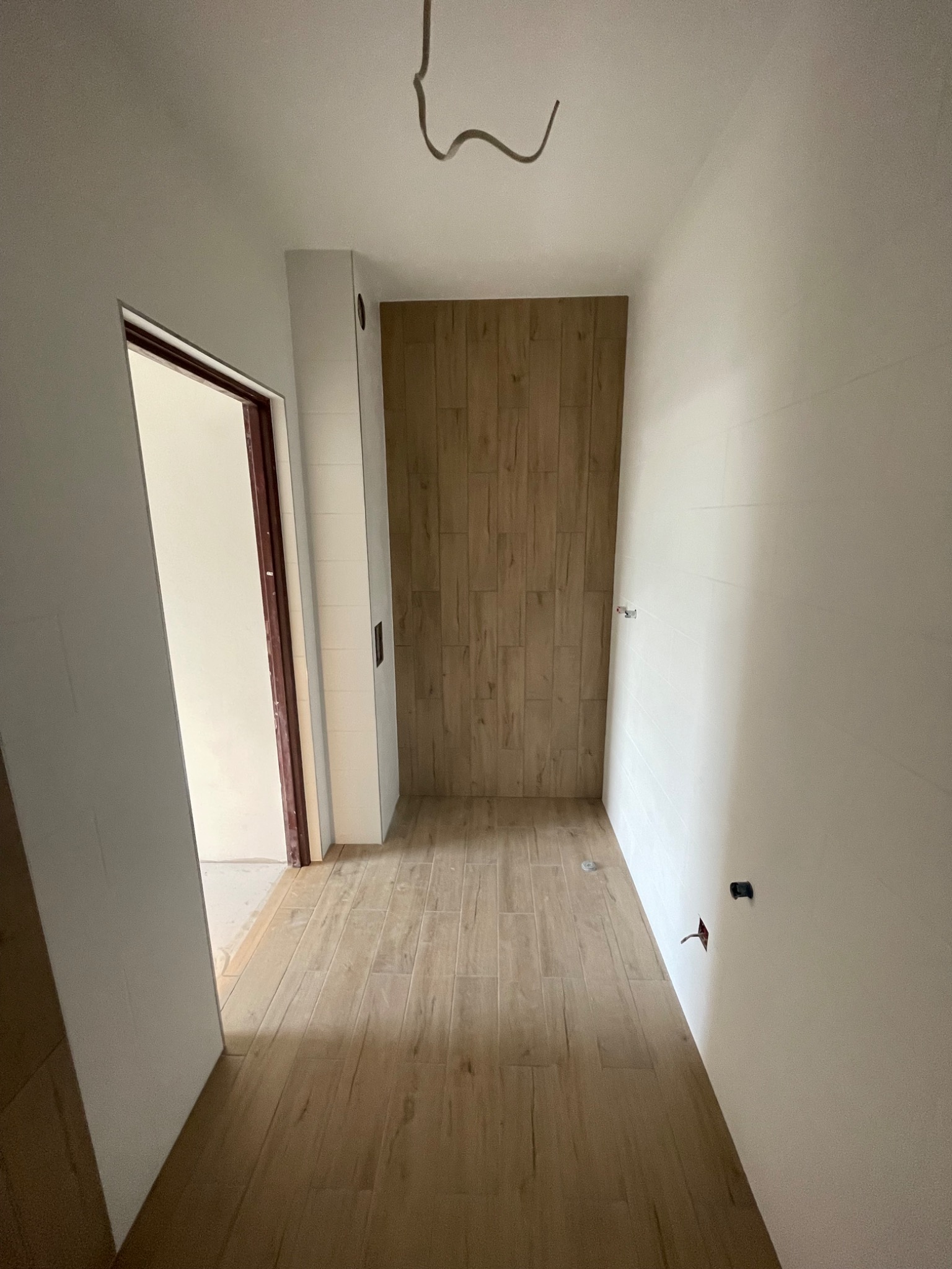 Wnętrze łazienki w trakcie wykańczania: drewnopodobne płytki na podłodze i ścianie prysznica, białe ściany, widoczne instalacje elektryczne, otwarte drzwi.