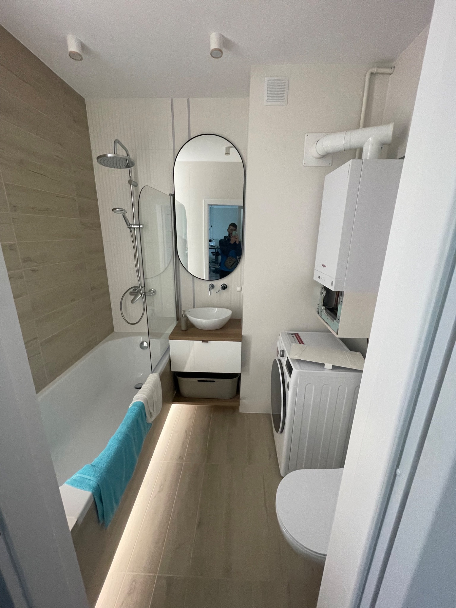 Nowoczesna łazienka z wanną, kabiną prysznicową, umywalką z szafką, pralką i toaletą. Beżowe płytki na ścianach i podłodze. Lustro owalne w czarnej ramie. Oświetlenie punktowe.