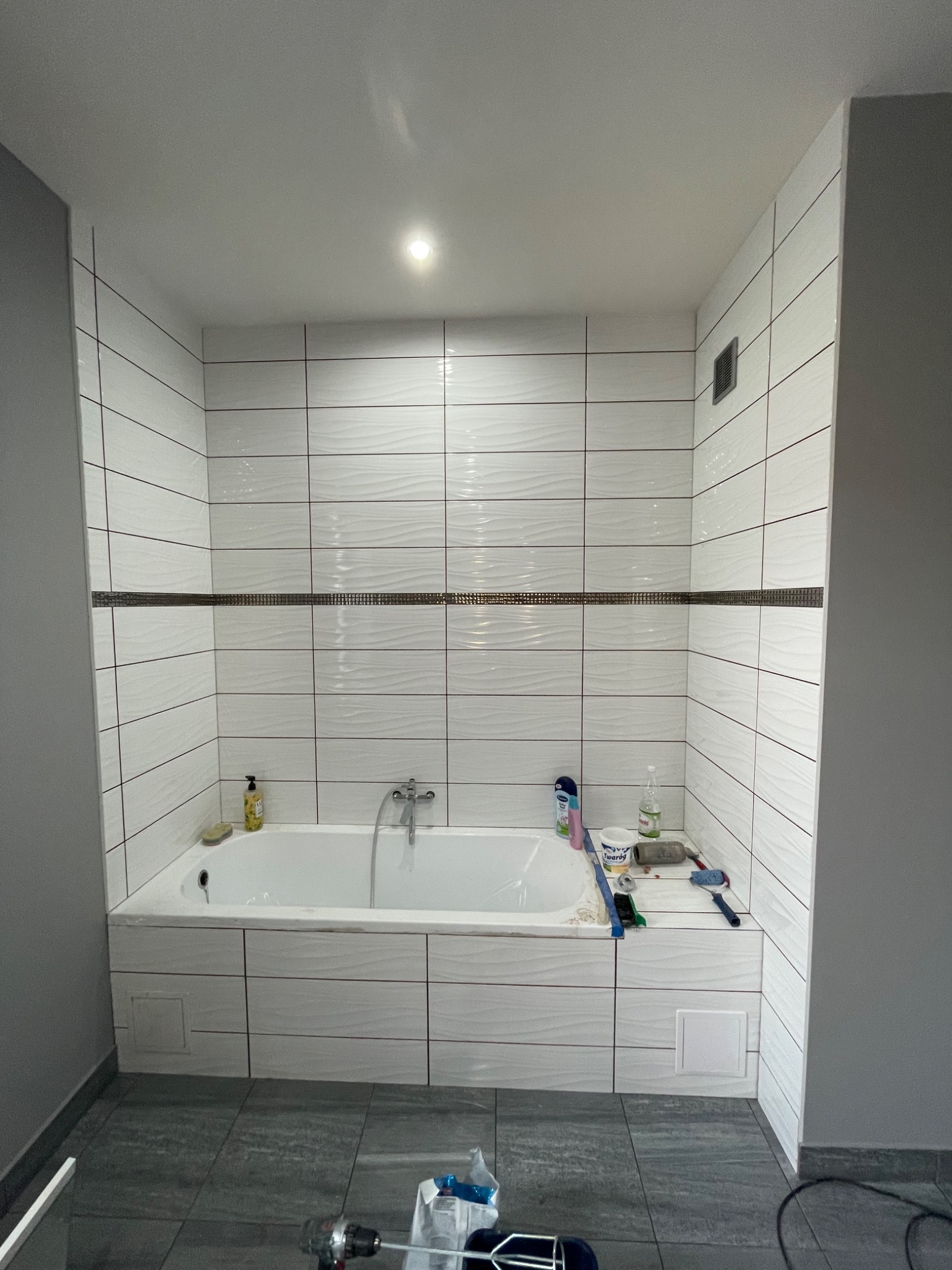 Wykończona łazienka z wanną obłożoną białymi, poziomymi płytkami z delikatnym wzorem fal. Ciemnoszara podłoga i fragment ściany. Widoczne narzędzia i akcesoria.