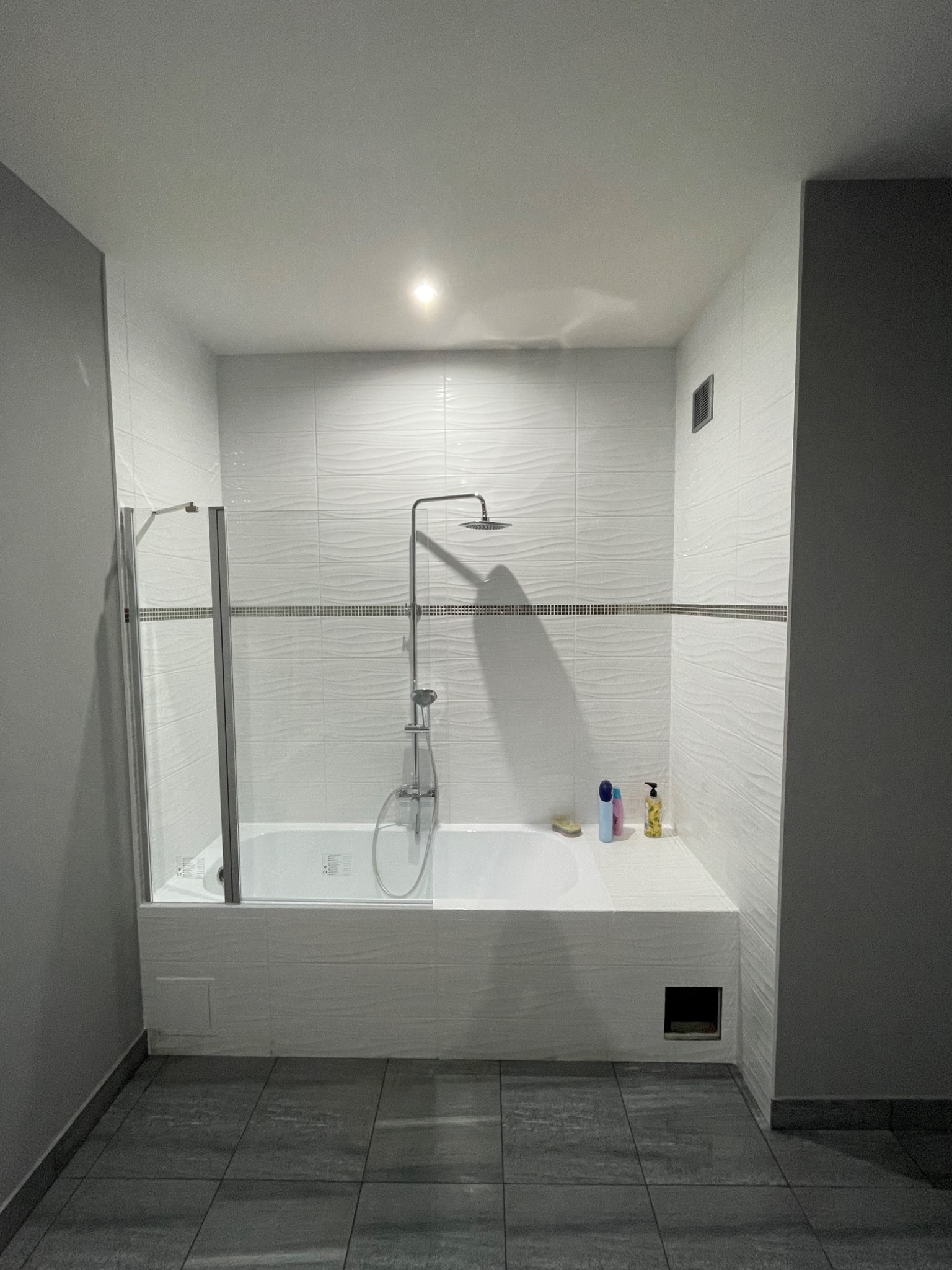 Nowoczesna łazienka po remoncie: białe płytki z falistym wzorem, szklany parawan nawannowy, szara podłoga. Minimalistyczny design z akcentem na teksturę.