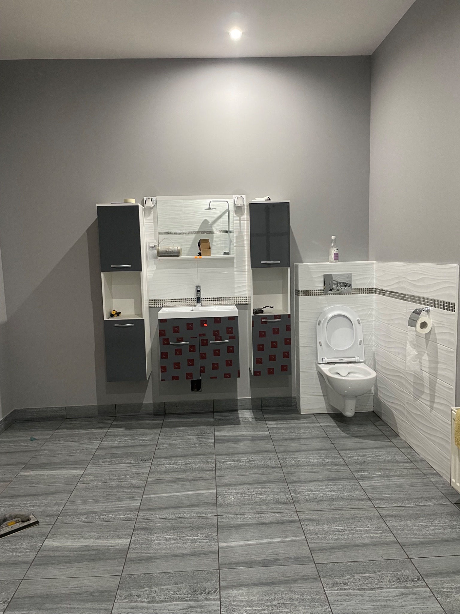 Wykończona łazienka w szarościach: płytki podłogowe imitujące drewno, białe płytki ścienne z delikatnym wzorem, szare szafki wiszące i toaleta. Widoczne narzędzia glazurnicze.