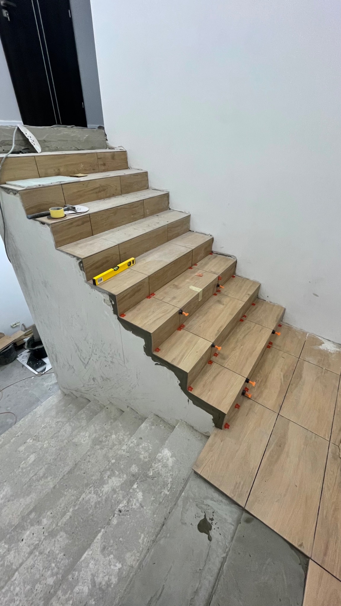 Układanie płytek imitujących drewno na schodach wewnątrz budynku. Widoczny poziomica, taśma malarska i system poziomowania płytek. Częściowo zakończone prace.