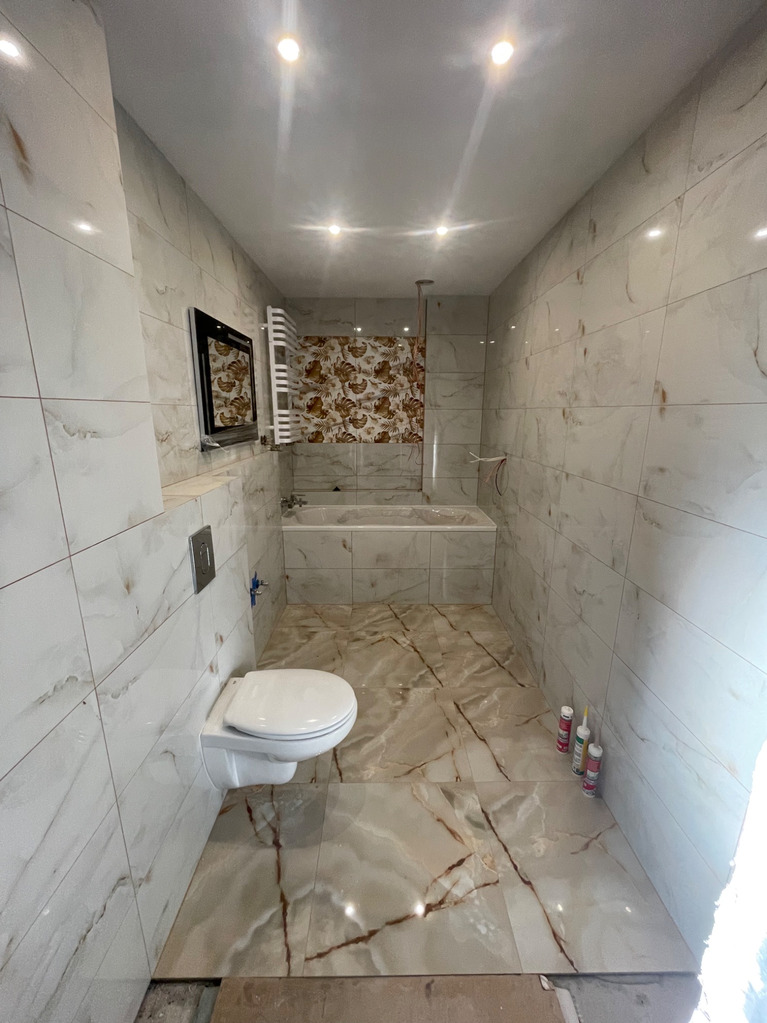 Wykończona łazienka z beżowymi płytkami imitującymi marmur na ścianach i podłodze. Wisząca toaleta, wanna i grzejnik drabinkowy. Widoczne silikony w rogu.