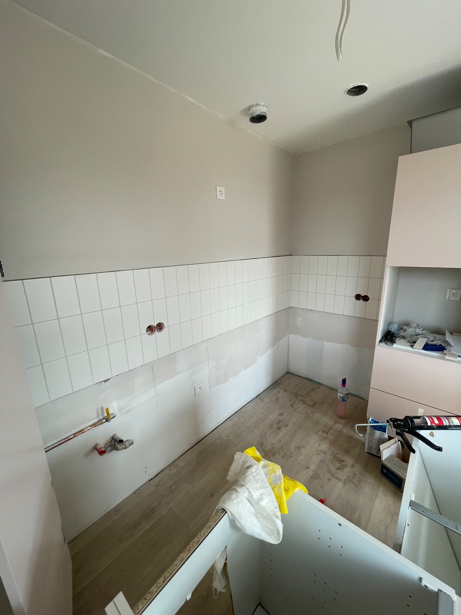 Wnętrze kuchni w trakcie remontu: częściowo ułożone białe płytki na ścianie, widoczne instalacje, niezabudowane szafki i akcesoria remontowe na podłodze.