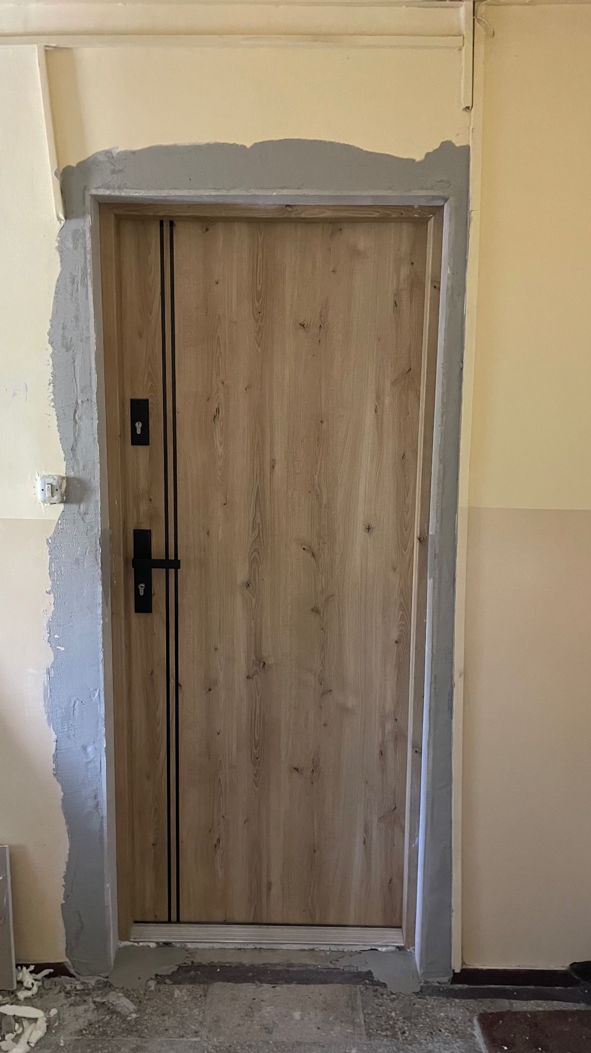 Zamontowane drzwi wewnętrzne z drewnianym fornirem i czarnymi akcentami, widoczne ślady po szpachlowaniu ościeżnicy, surowy stan wykończenia.