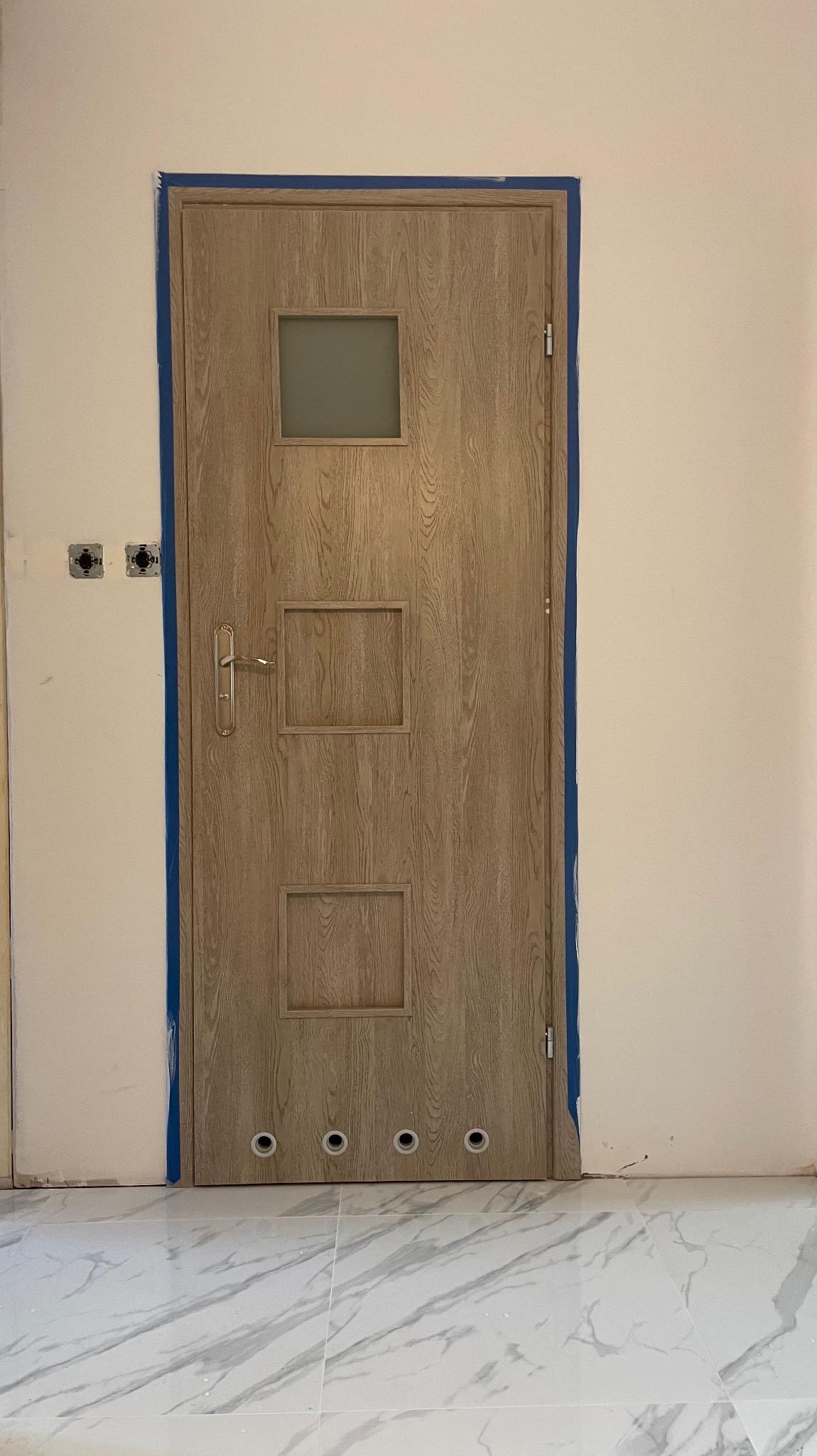 Jasne, drewnopodobne drzwi z szybką i otworami wentylacyjnymi, zabezpieczone niebieską taśmą malarską, na tle białej ściany i marmurowej podłogi.