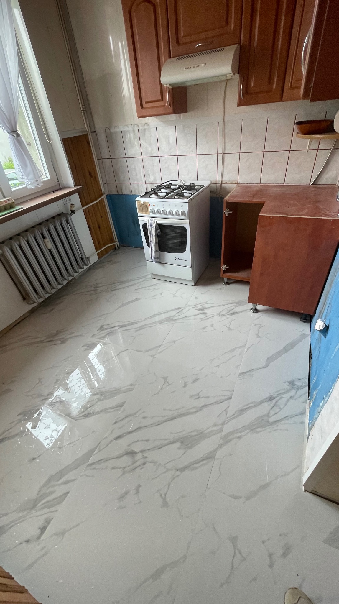 Jasne, marmurowe płytki podłogowe w kuchni, z widoczną kuchenką i fragmentami szafek. Ujęcie z perspektywy podłogi, podkreślające wzór płytek.