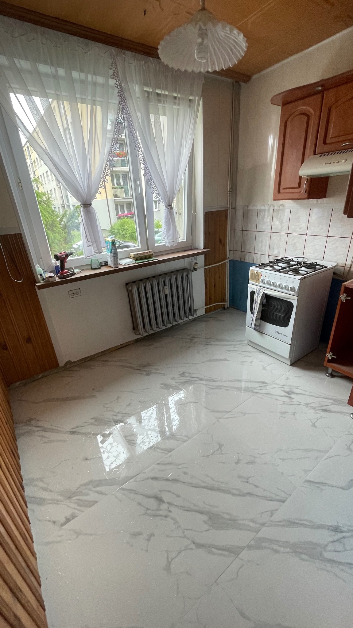 Kuchnia po remoncie: białe płytki imitujące marmur na podłodze, okno z firankami, grzejnik, kuchenka. Widoczne narzędzia na parapecie.