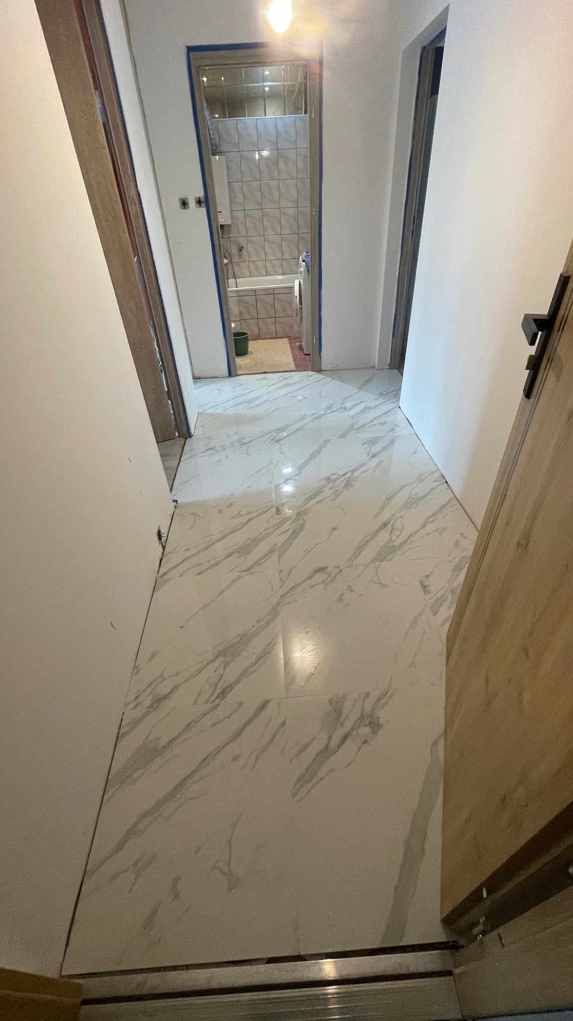Położone białe płytki imitujące marmur na podłodze w korytarzu, widok na łazienkę z wanną i pralką. Świeżo wykończone wnętrze z otwartymi drzwiami.
