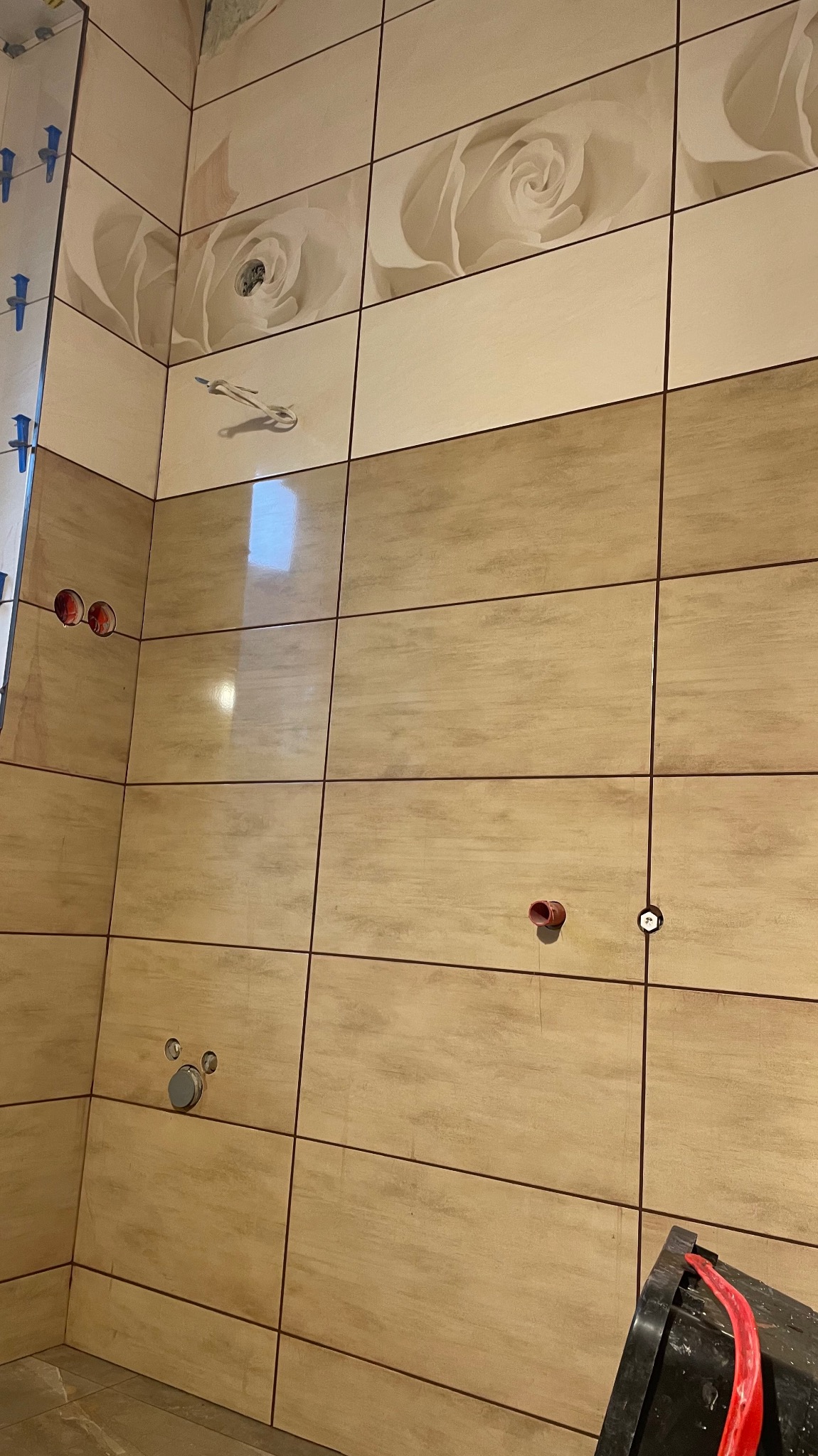 Ściana łazienki wyłożona beżowymi płytkami z motywem kwiatowym w górnej części. Widoczne otwory na instalacje i narzędzia glazurnicze.