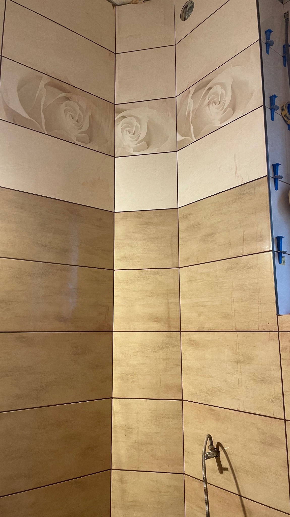 Wykończona ściana łazienki z beżowych płytek z motywem kwiatowym, widoczne plastikowe krzyżyki glazurnicze i fragment baterii. Ukończona realizacja glazurnicza.