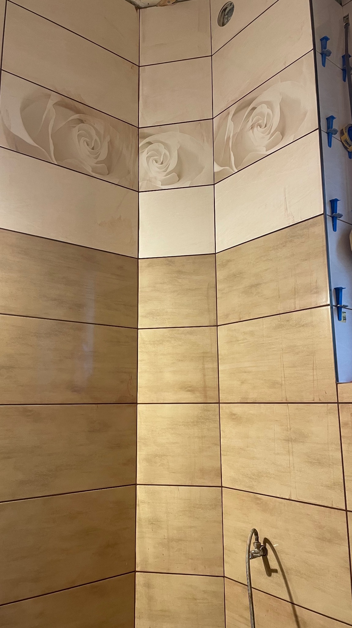 Wykończona ściana w łazience w Piekarach Śląskich, z beżowymi płytkami z motywem kwiatowym. Widoczne fugi i fragment instalacji wodnej.