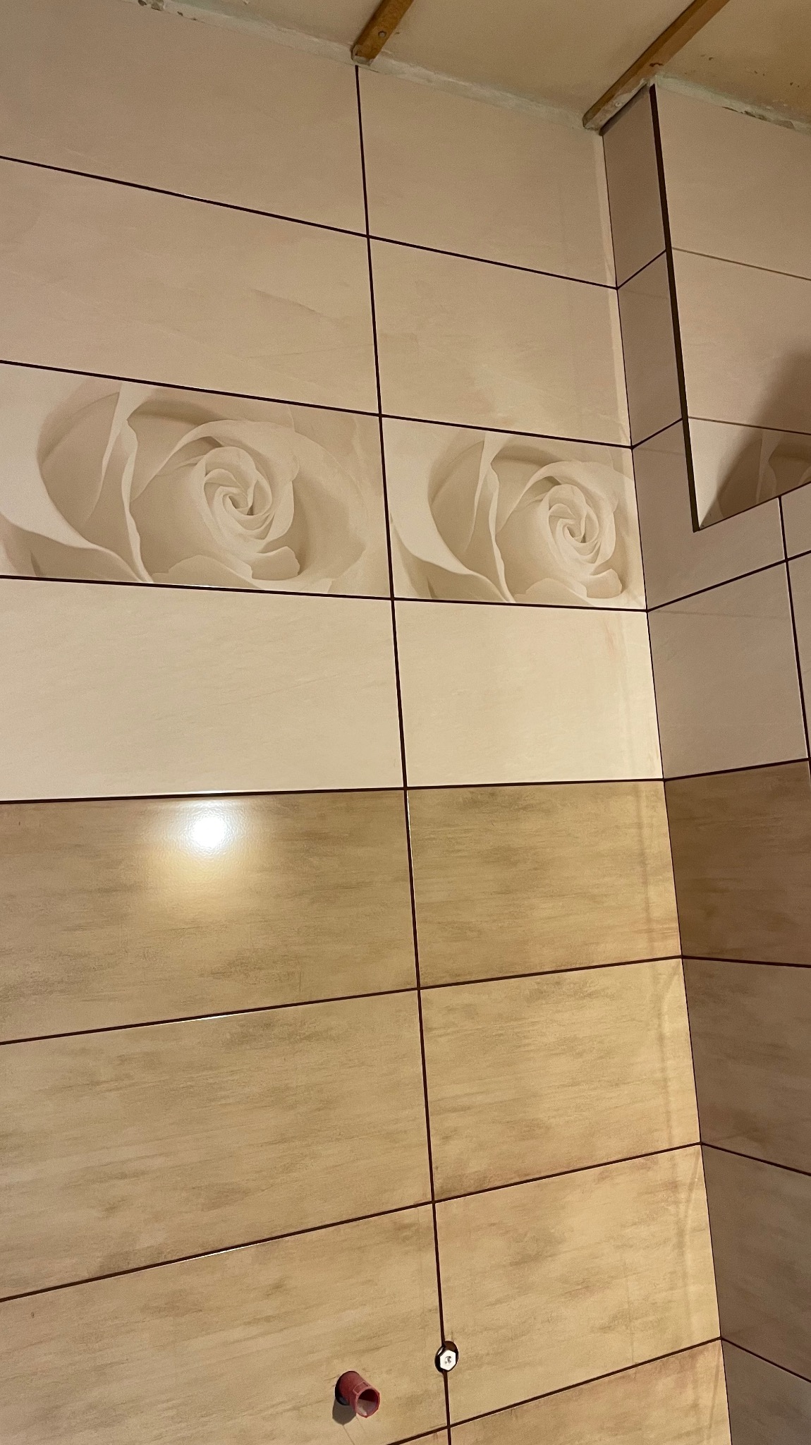 Beżowe, prostokątne płytki z motywem róży na ścianie łazienki, widoczne fragmenty instalacji wodnej. Ukończone wykończenie ściany w neutralnej kolorystyce.