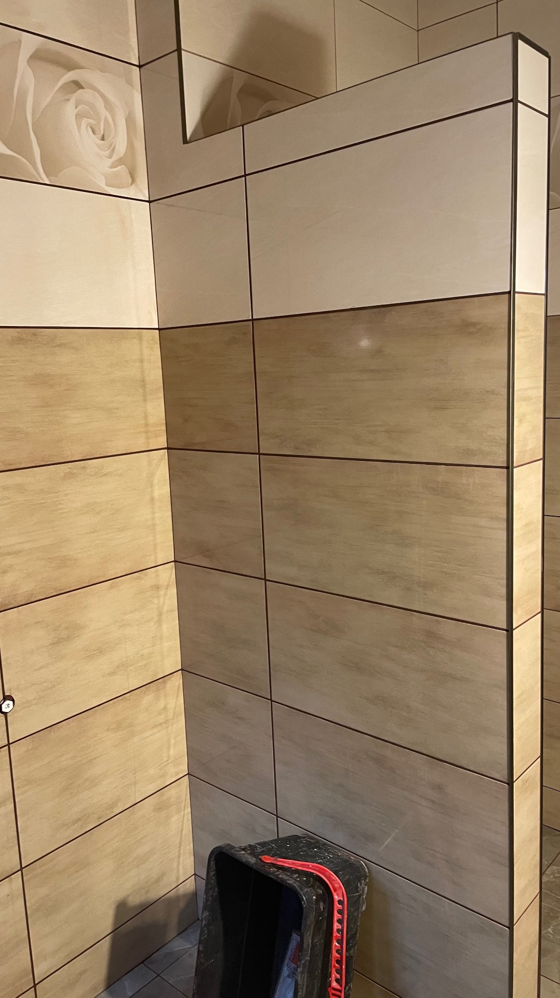 Wykończona ściana w łazience z beżowymi i kremowymi płytkami. Na górze płytka z motywem kwiatowym. W rogu stoi czarne wiadro z czerwoną rączką.