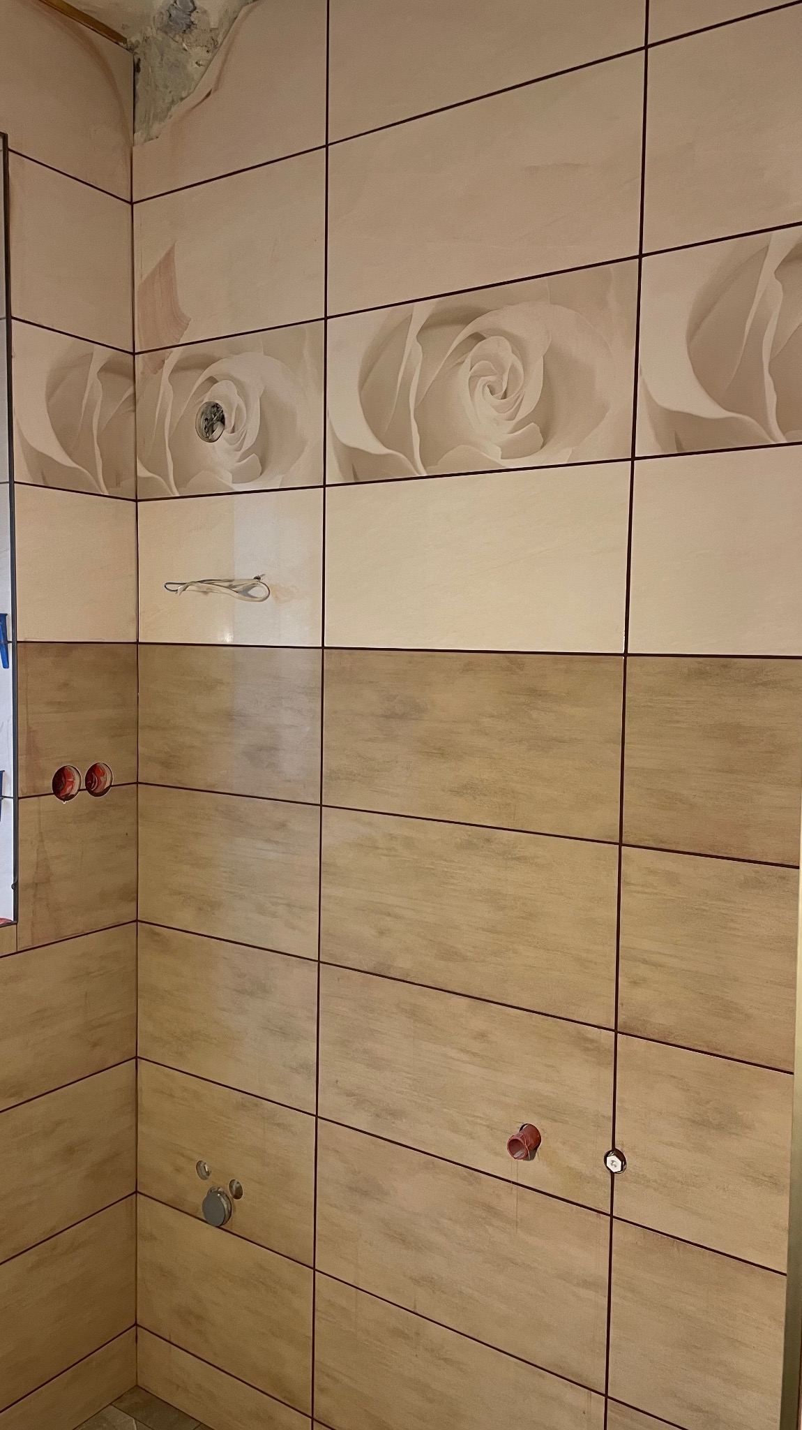 Wykończona ściana łazienki z beżowymi płytkami, dekorowana motywem kwiatowym w górnej części. Widoczne otwory na armaturę i fragment sufitu z ubytkiem tynku.