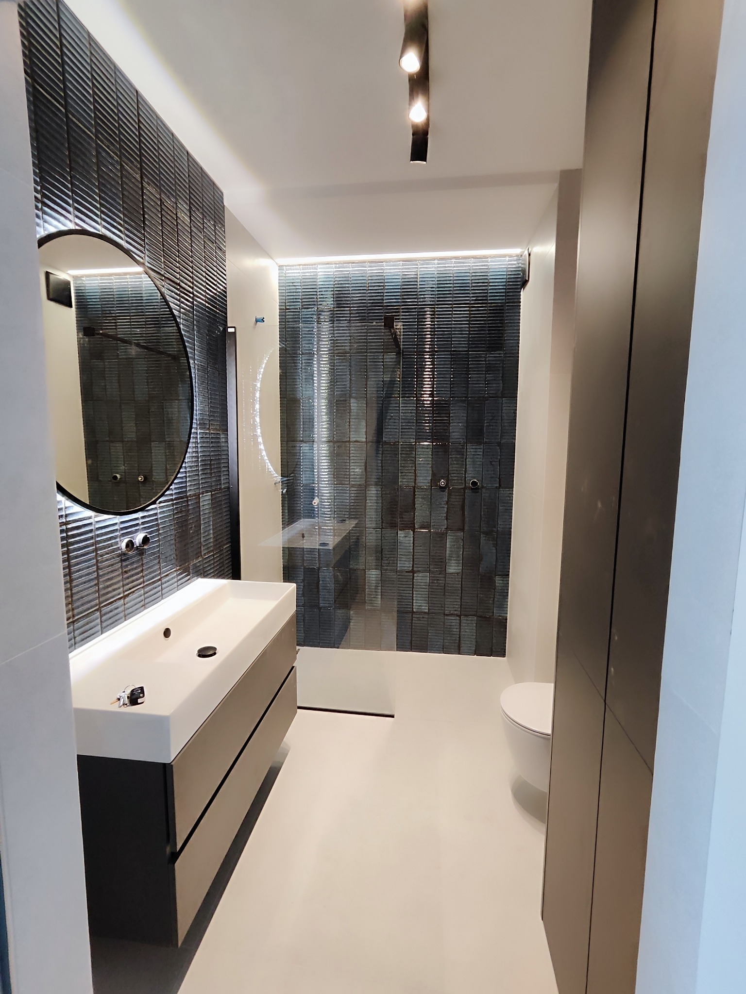 Nowoczesna łazienka z ciemnymi, teksturowanymi płytkami, okrągłym lustrem i białą umywalką, oświetlona punktowo, z kabiną prysznicową typu walk-in.