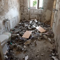 ProPerfect - Łazienka w trakcie demontażu: gruz, odłupane płytki, odsłonięte ściany. Widoczne fragmenty toalety i okno z zielenią na zewnątrz. Stan przed remontem.