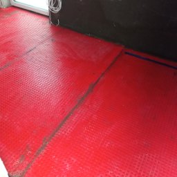 ProPerfect - Czerwona mata grzewcza podłogowa z wypustkami, ułożona w rogu pomieszczenia z widocznymi kablami i fragmentem ściany przygotowanej do dalszych prac wykończeniowych.