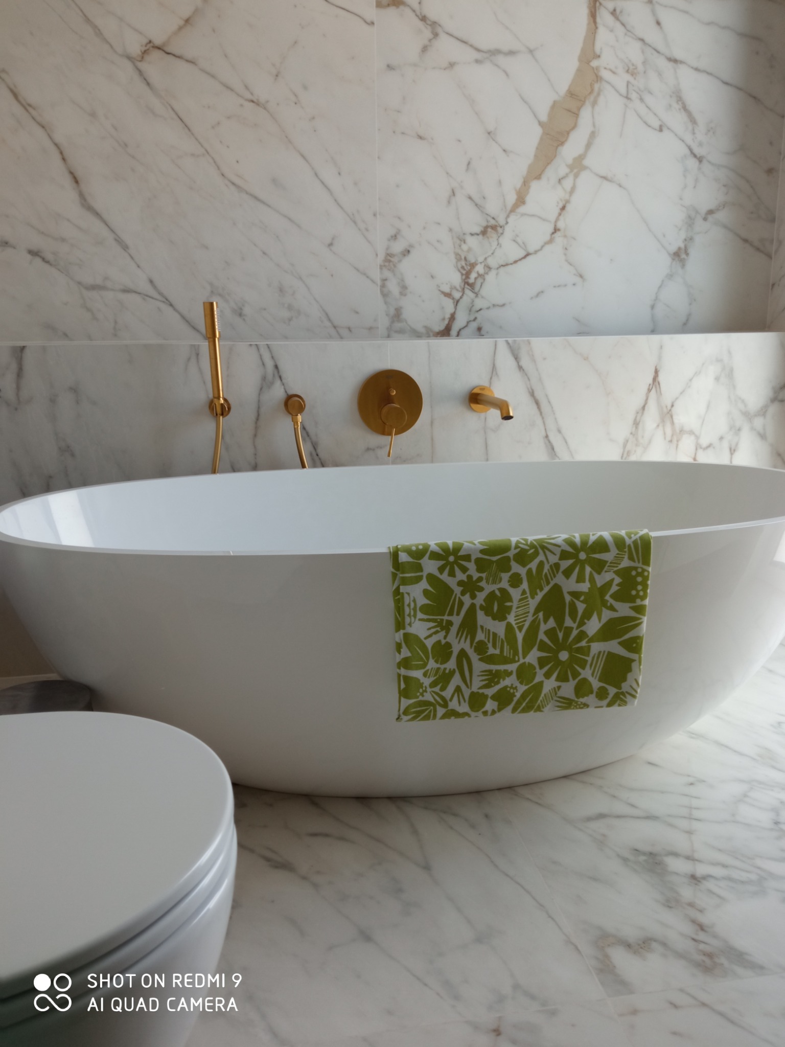 Nowoczesna łazienka z białą wanną wolnostojącą, złotą armaturą i marmurowymi płytkami. Na wannie leży zielony ręcznik w kwiaty. Widoczny fragment toalety.